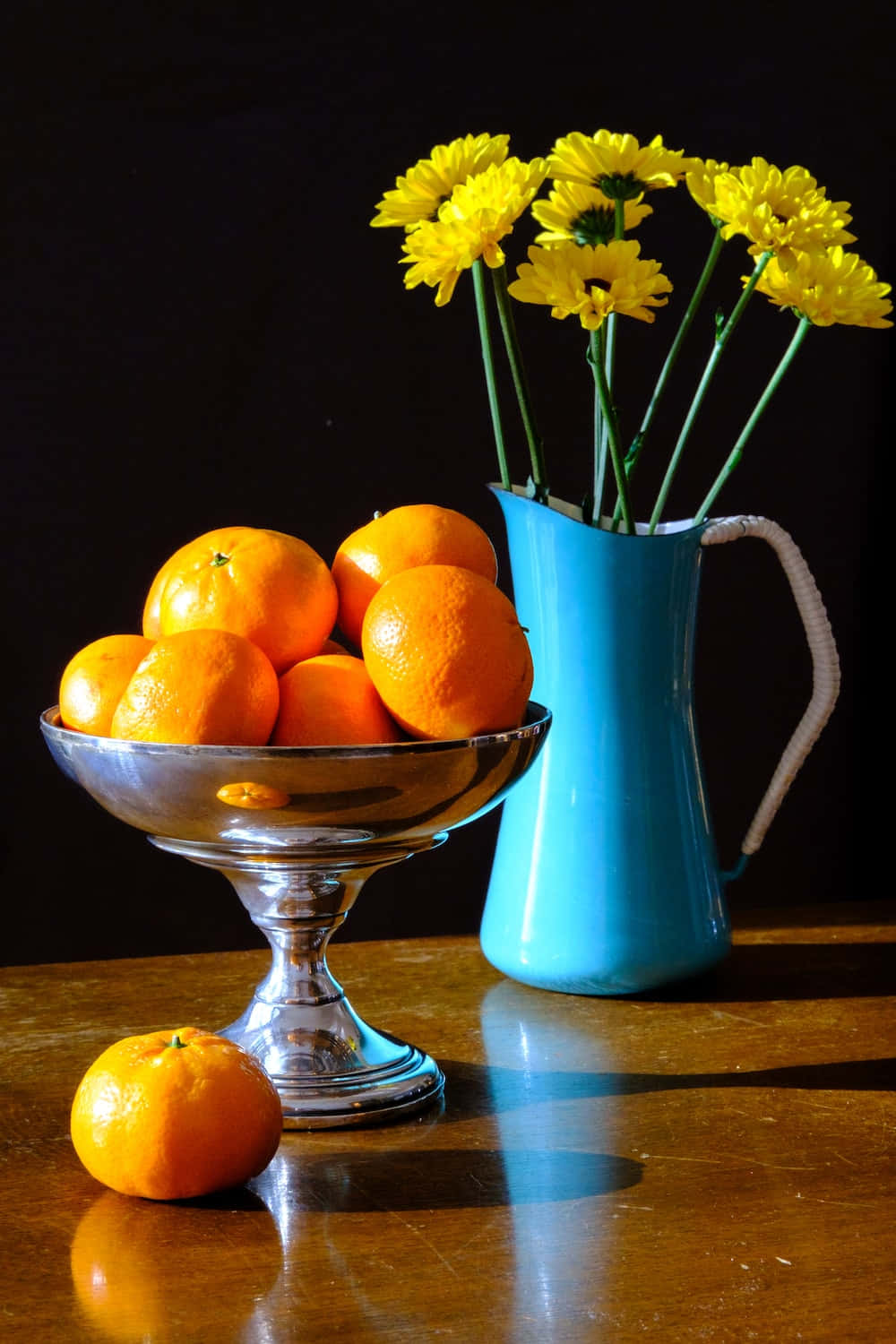 still life 桌布 壁紙 — 一個銀碗盛著鮮豔的柳橙，放在一張拋光木桌上，旁邊是一個藍色水壺，插著明亮的黃色雛菊。