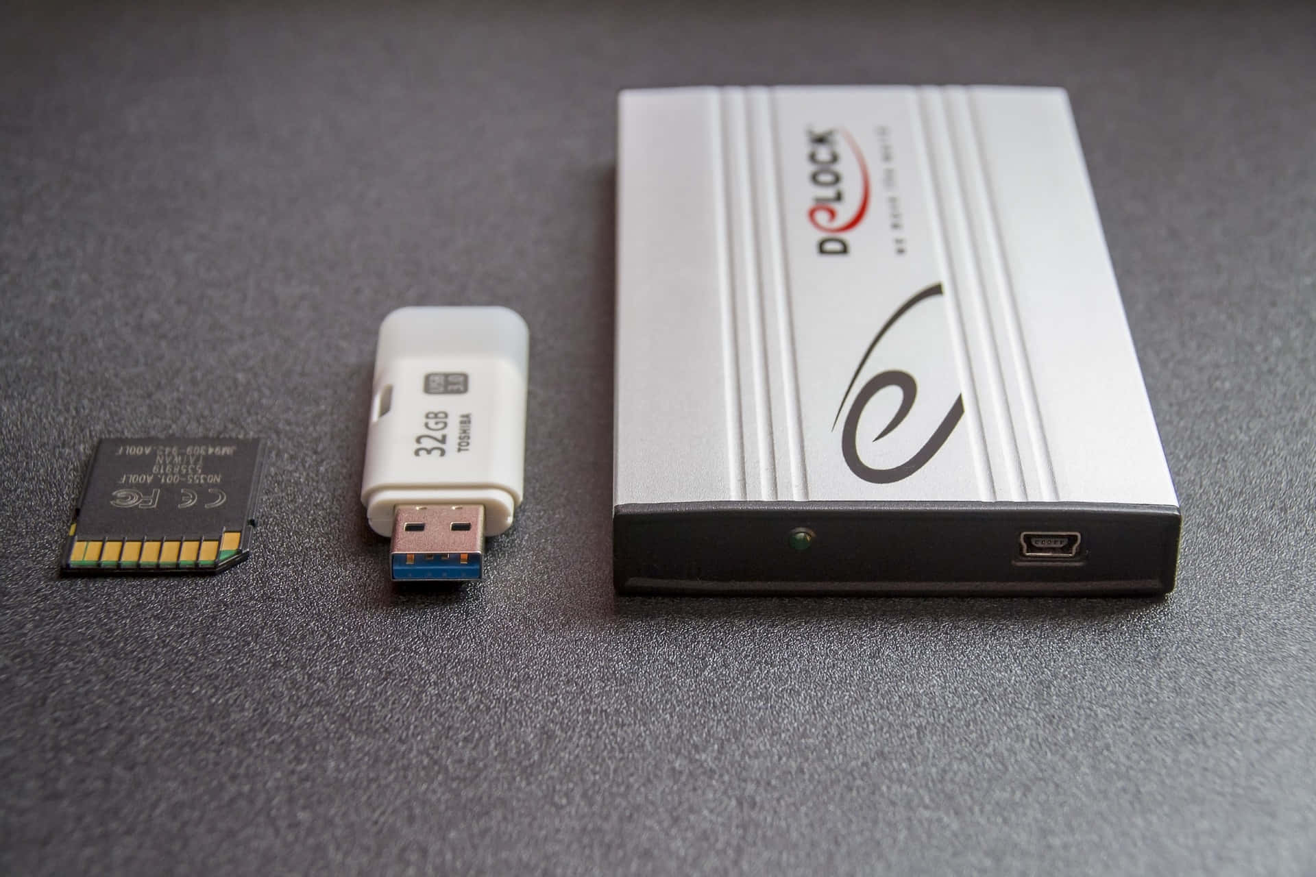 Backup 桌布 壁紙 — 一個銀色 D-LOCK 外接硬碟旁邊放著一個白色 32GB USB 隨身碟和一個黑色 SD 卡，置於灰色紋理表面上。