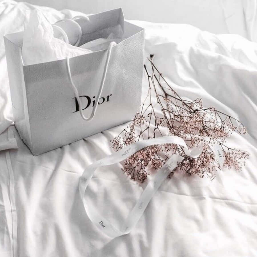 Dior 動態 桌布 壁紙 — 一個銀色Dior購物袋放在皺巴巴的白色床單上，旁邊有乾燥的粉紅色花朵和相配的絲帶。