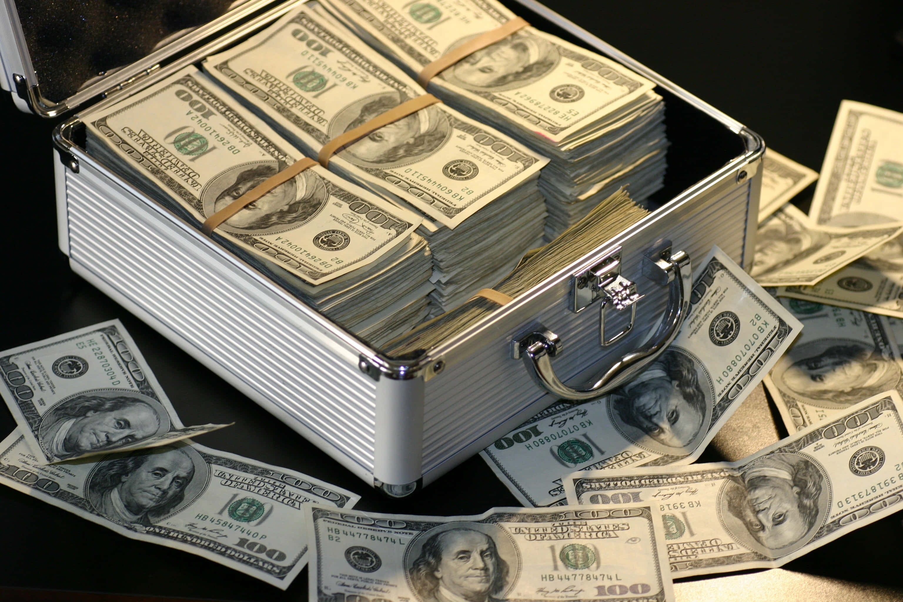 金錢 桌布 壁紙 — 一個銀色公事包裝滿一疊疊百元美鈔，將現金灑在深色表面上。