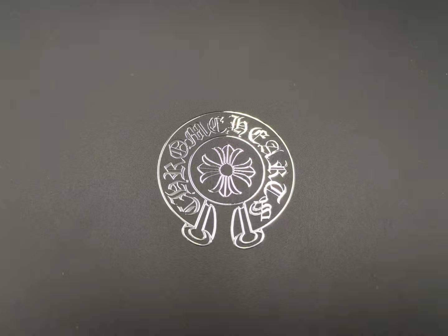 Chrome Hearts 桌布 壁紙 — 一個銀色圓形徽章，帶有華麗的哥德式字體和中央花卉圖案，放置在深灰色紋理表面上。