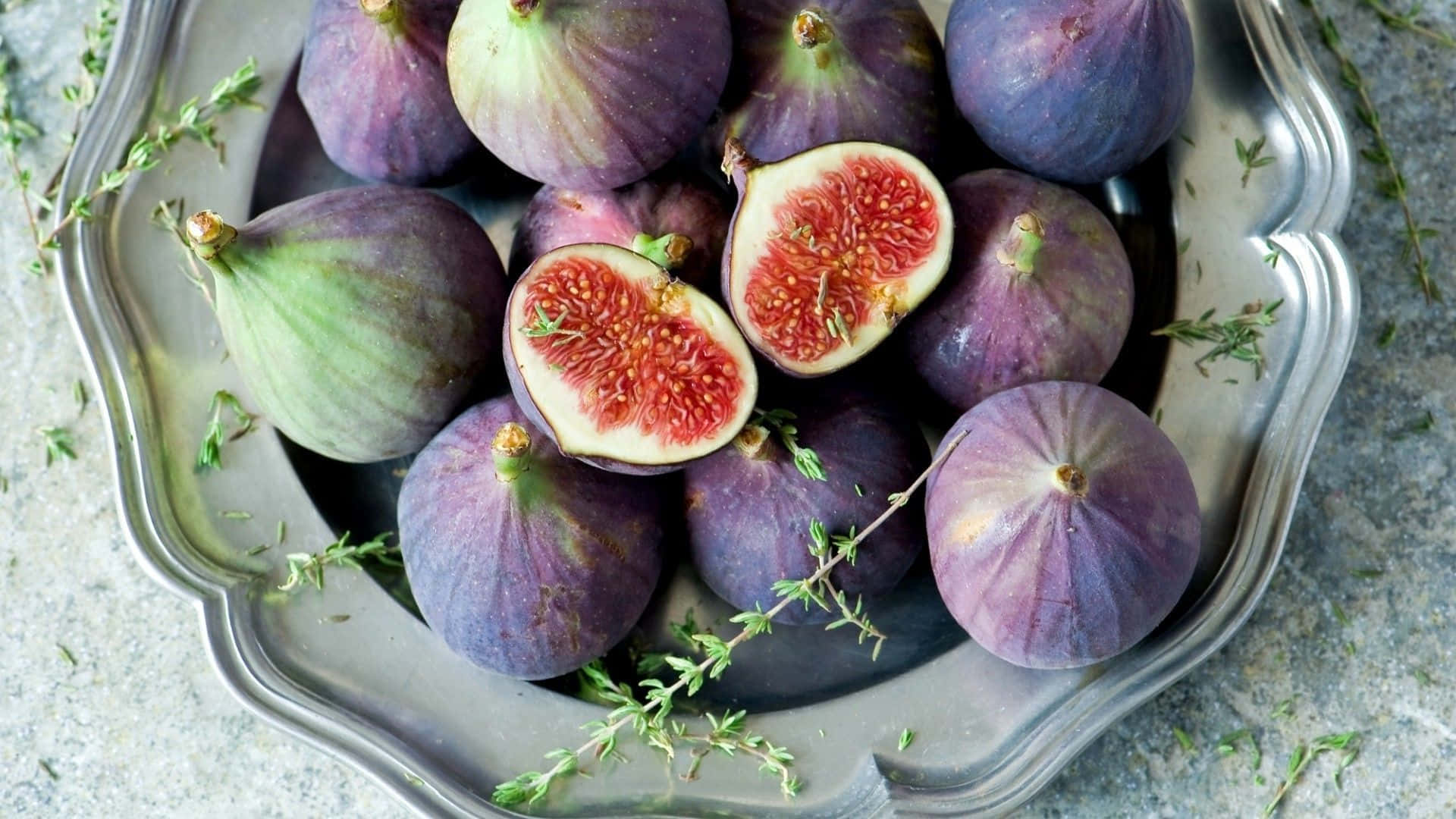 Purple Figs 桌布 壁紙 — 一個銀色扇貝形盤子盛放著成熟的紫色無花果，其中兩個切開露出粉紅色果肉，並有綠色百里香枝散落周圍。