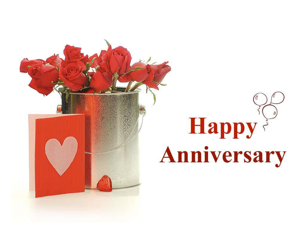 Happy Anniversary 桌布 壁紙 — 一個銀色桶裝滿鮮豔的紅玫瑰，旁邊放著一張紅色心形卡和一個帶有「Happy Anniversary」文字和氣球塗鴉的巧克力糖果。