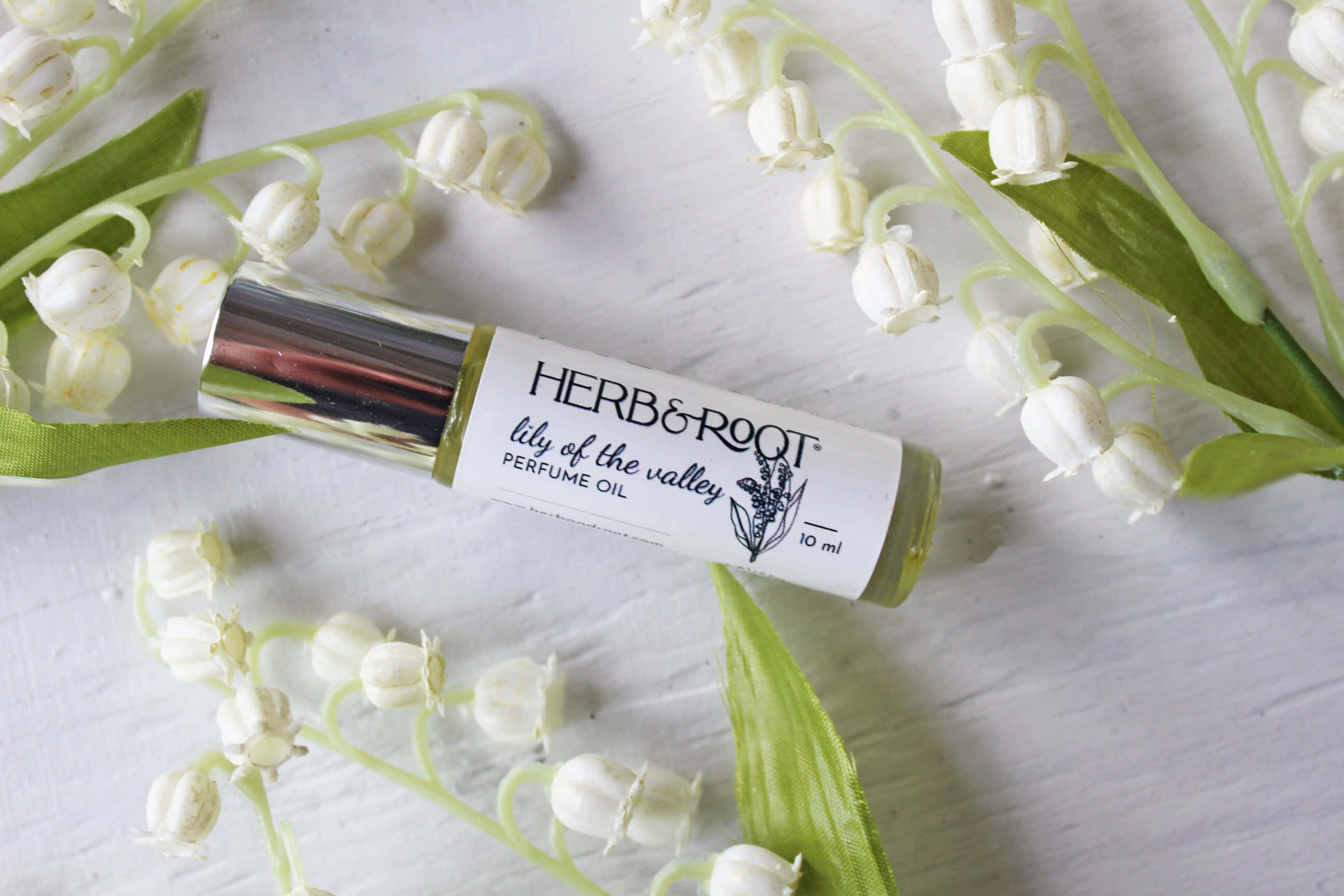 Floral Fragrance 桌布 壁紙 — 一個銀色滾珠香水油，標籤為鈴蘭，放在有紋理的白色表面上，周圍是新鮮的白色鐘形花朵。