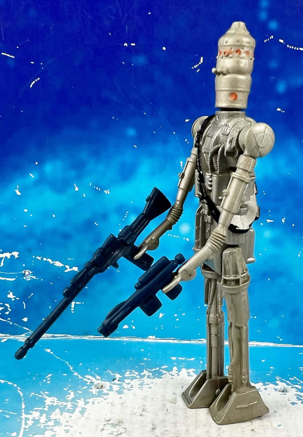 Ig-88 桌布 壁紙 — 一個銀色玩具機器人站立著，在帶有白色潑濺的紋理藍色背景前，手持兩把黑色爆能槍。
