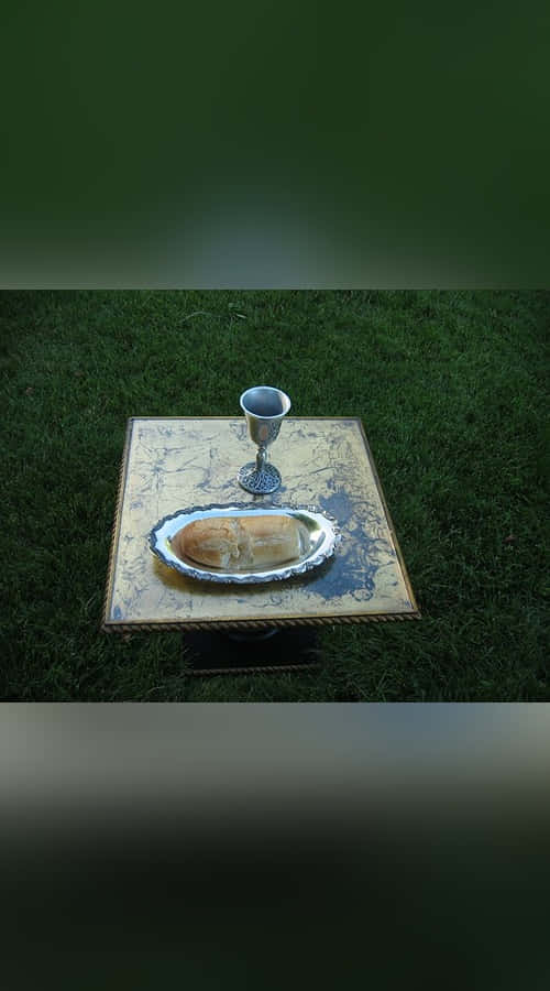 Sacrament 桌布 壁紙 — 一個銀色聖杯和麵包放在一個華麗的托盤上，擱在一張放在茂密綠色草坪上的小桌子上。
