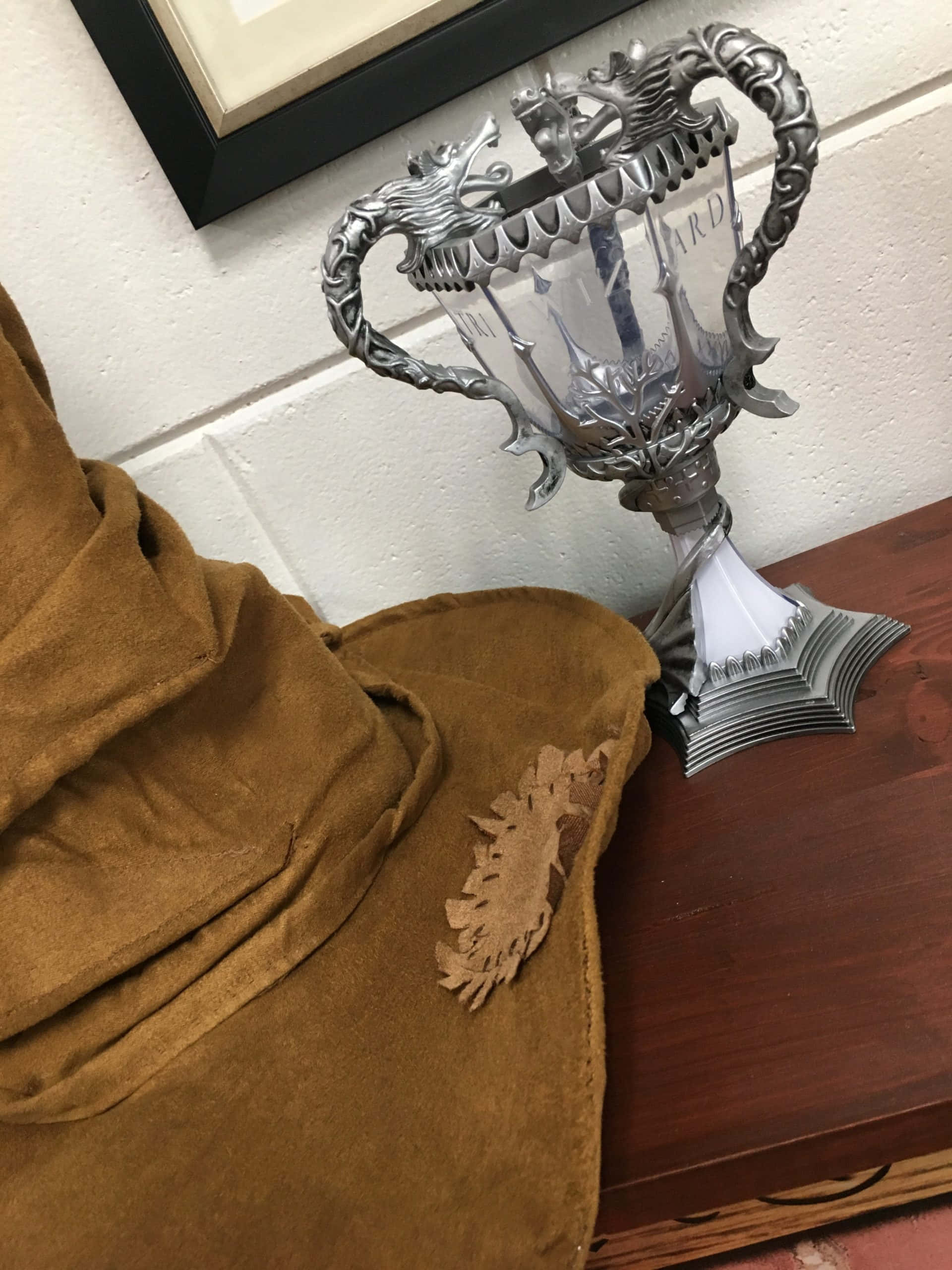 The Triwizard Cup 桌布 壁紙 — 一個銀色龍柄獎盃放在木質表面上，旁邊有一張棕色麂皮毯子，背景是白色磚牆。