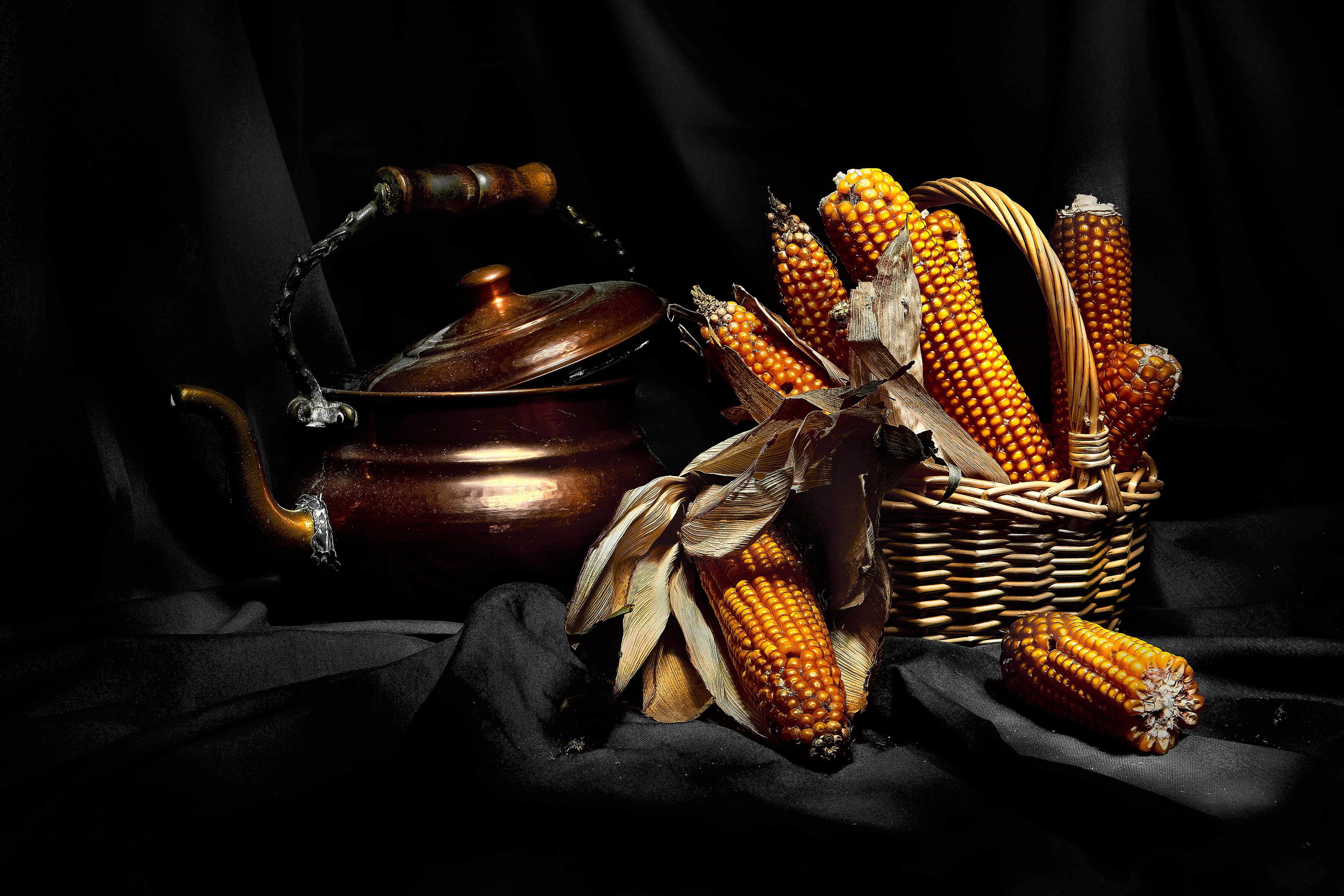 Corn 桌布 壁紙 — 一個銅壺和一籃金黃色的玉米放在深色布料上，在戲劇性的燈光下。