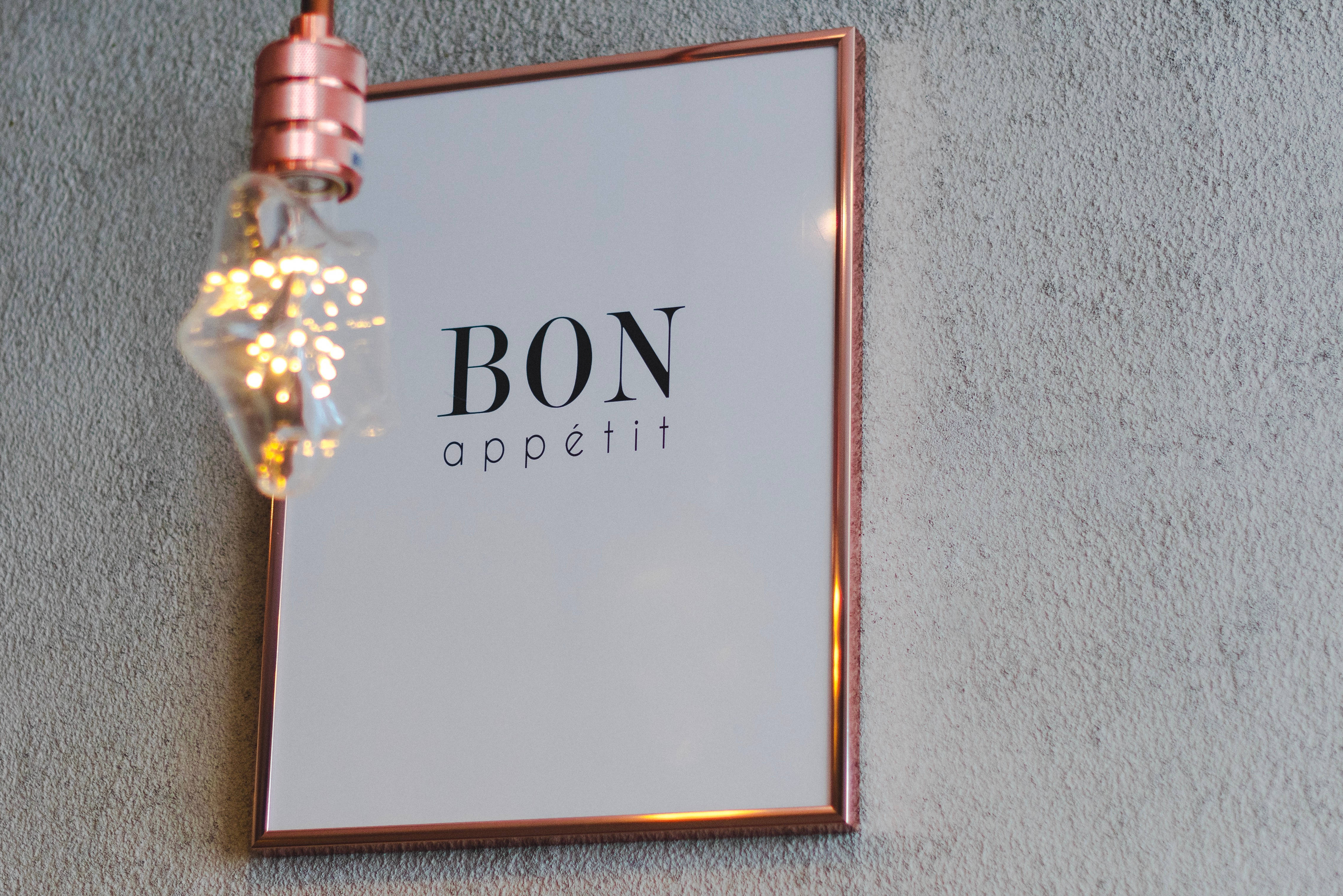 玫瑰金 桌布 壁紙 — 一個銅框 'Bon Appétit' 標誌掛在紋理灰色牆上，旁邊有一個發光的復古燈泡。