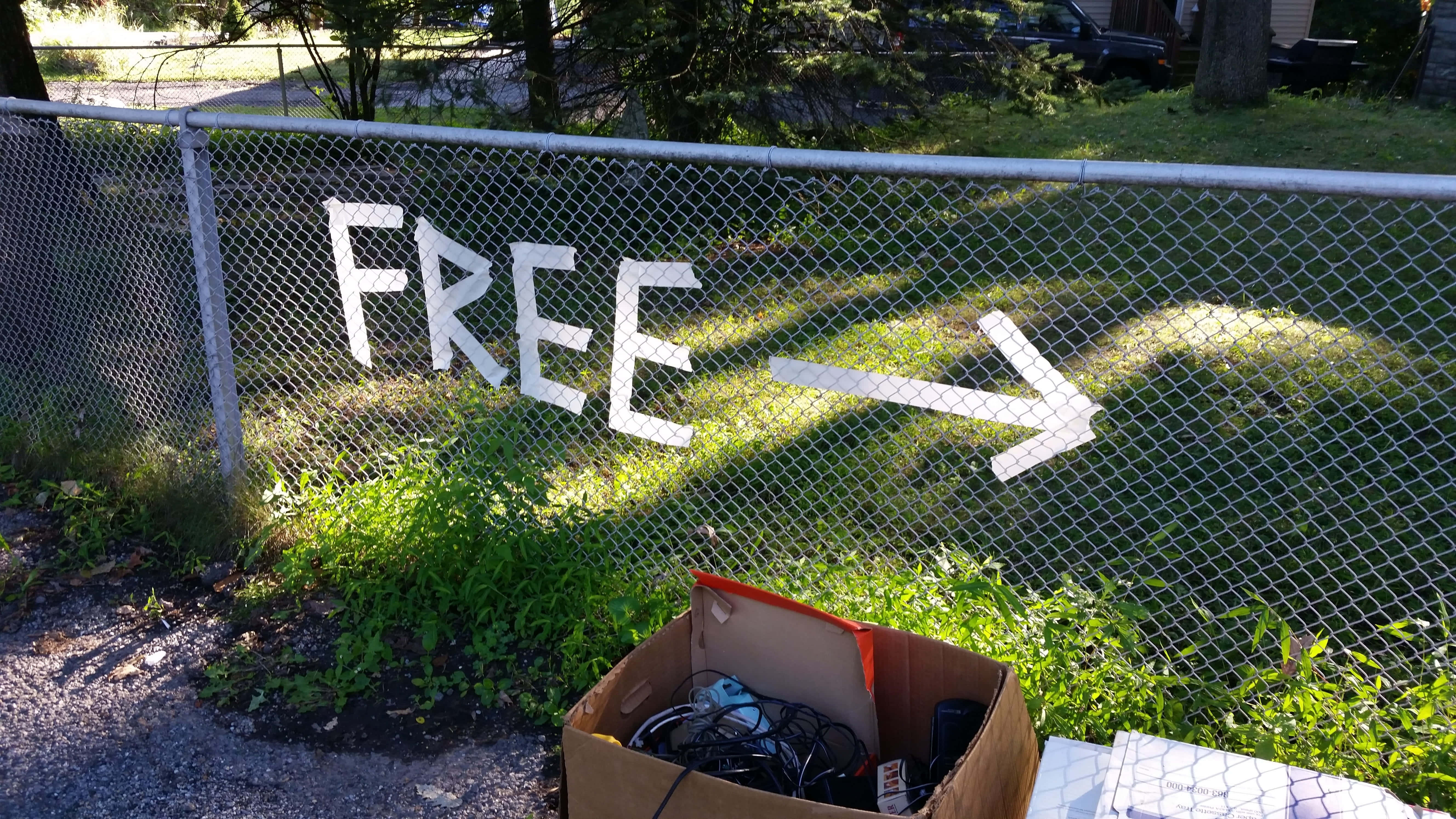 Free 桌布 壁紙 — 一個鐵絲網圍欄上畫有「FREE」和一個箭頭，指向一個放在草地上的紙箱，裡面裝滿了廢棄物品。
