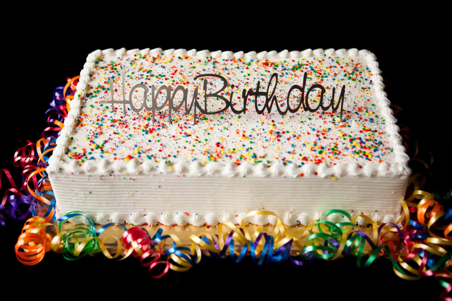 Happy Birthday Cake 桌布 壁紙 — 一個長方形生日蛋糕，上面有白色糖霜和彩色糖粒，周圍環繞著鮮豔的彩帶，背景為黑色。