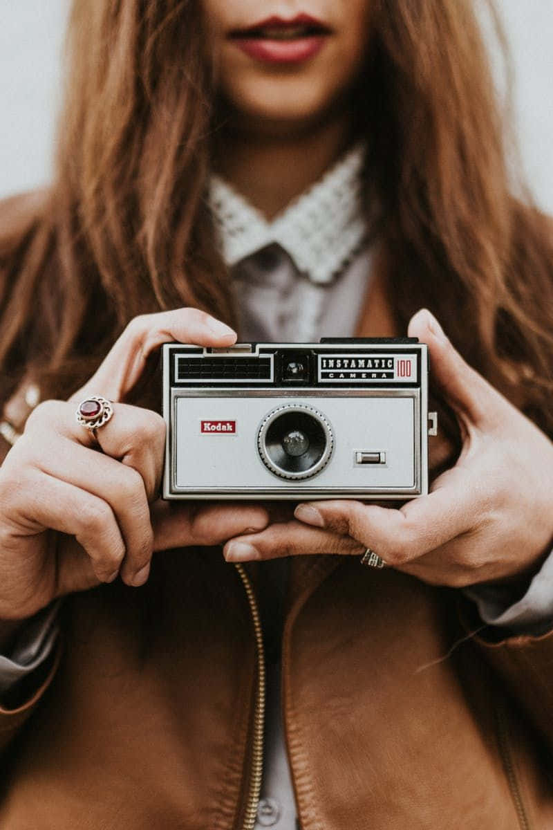 Holding Camera 桌布 壁紙 — 一個長棕色頭髮的人雙手拿著一部復古銀色Kodak Instamatic相機，背景柔和模糊。