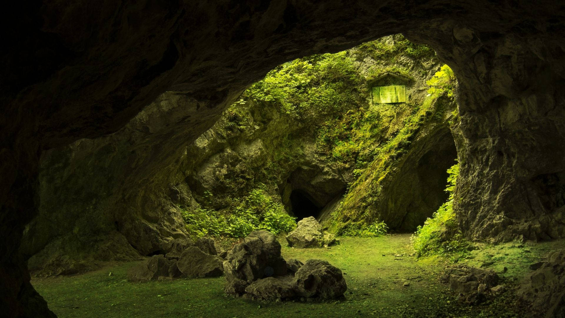 Cave 桌布 壁紙 — 一個長滿苔蘚的洞穴入口，框住了一個陽光明媚的叢林場景，有充滿活力的綠色樹葉和一個藏在岩石中的小木結構。