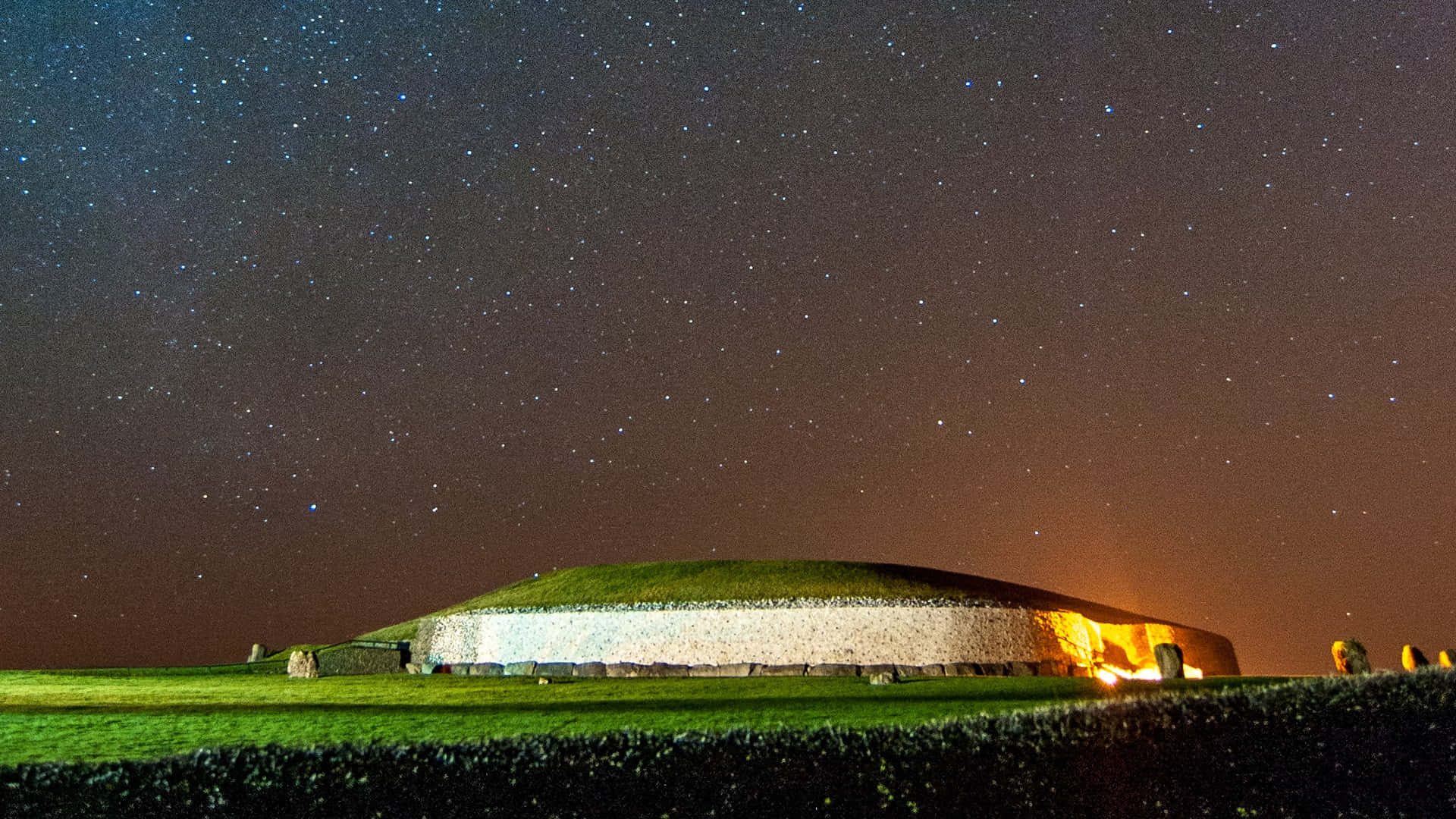 Stars 桌布 壁紙 — 一個長滿草的古墓在繁星點點的夜空下溫暖地發光，天空從深藍色漸變到地平線附近的橙色。