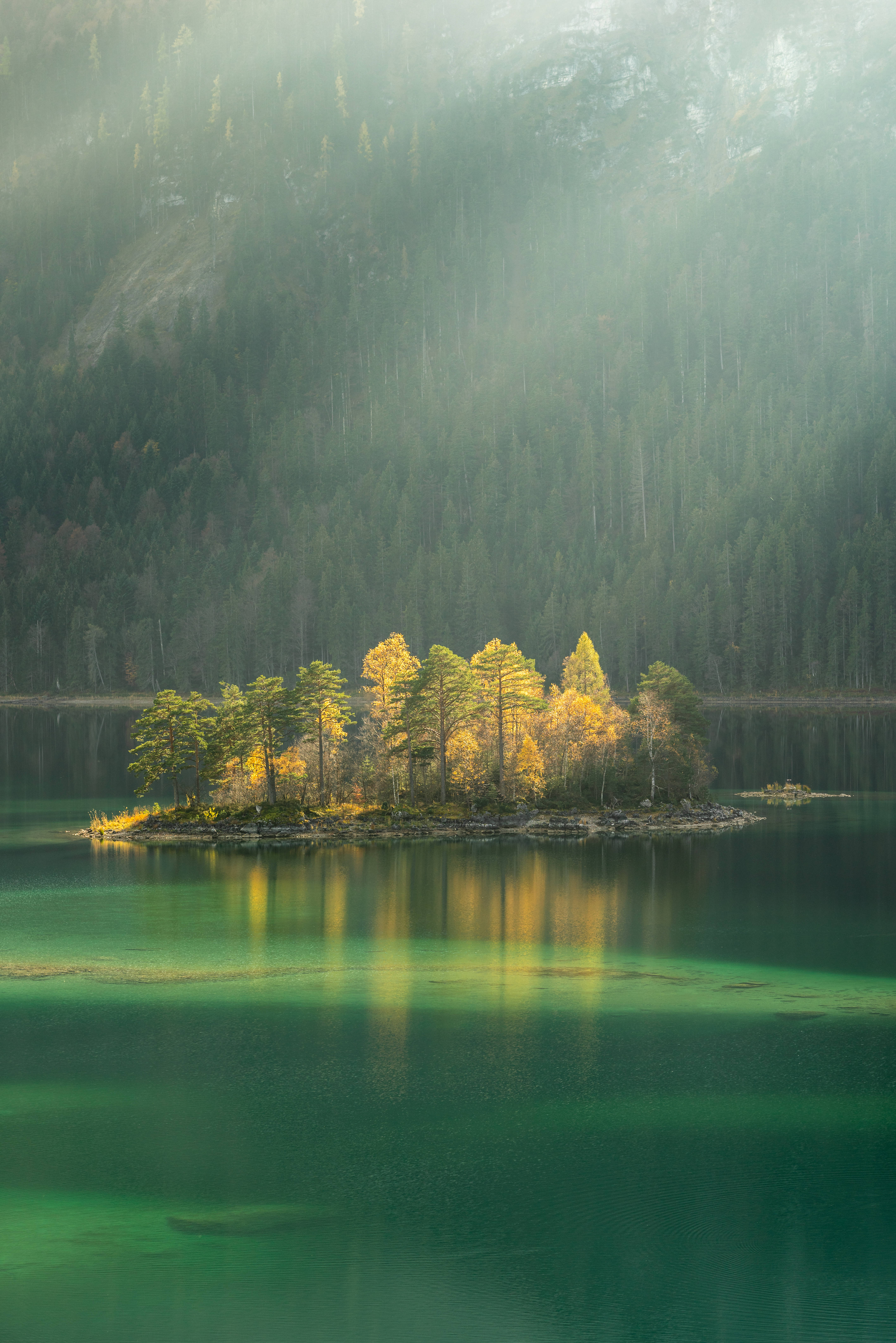 Lake 桌布 壁紙 — 一個長滿金色秋樹的小森林島嶼，坐落在霧氣瀰漫的翡翠湖中，四周環繞著茂密的常綠山脈。