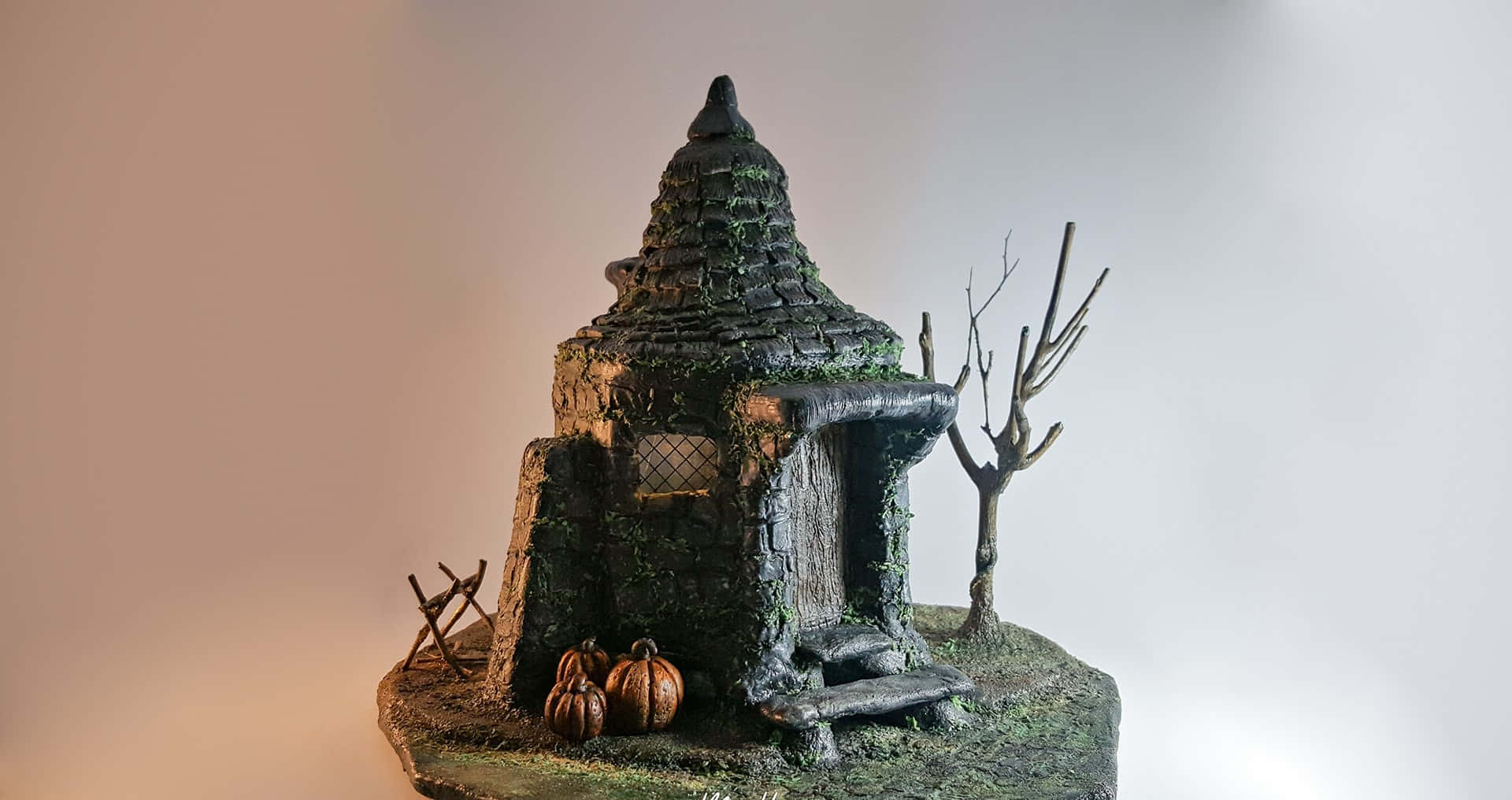 Hagrid's Hut 桌布 壁紙 — 一個長滿青苔的迷你鬼屋，放在岩石底座上，配有南瓜和一棵光禿禿的樹，背景是柔和的漸層色。