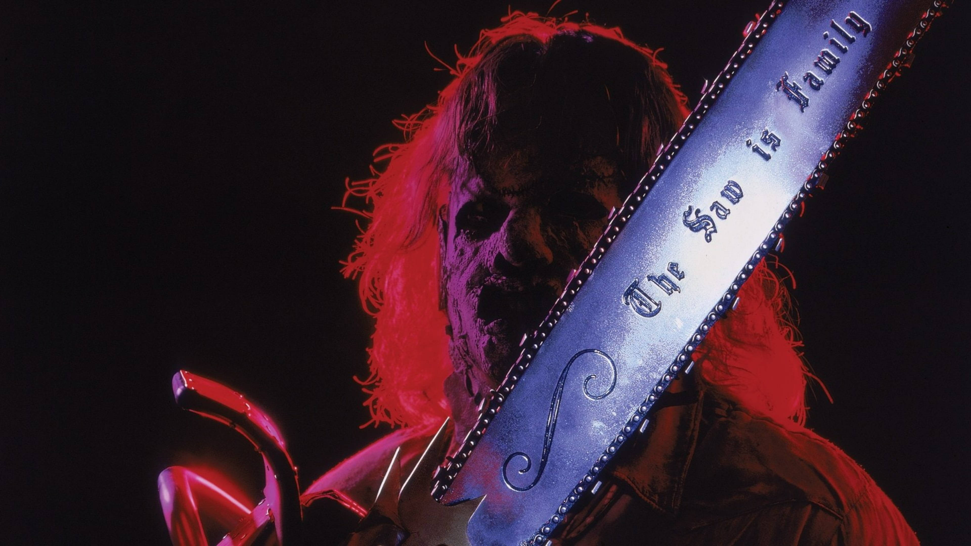 Leatherface 桌布 壁紙 — 一個長紅頭髮的威脅人物，在戲劇性的紅光下拿著一把刻有「The Saw is Family」的電鋸。