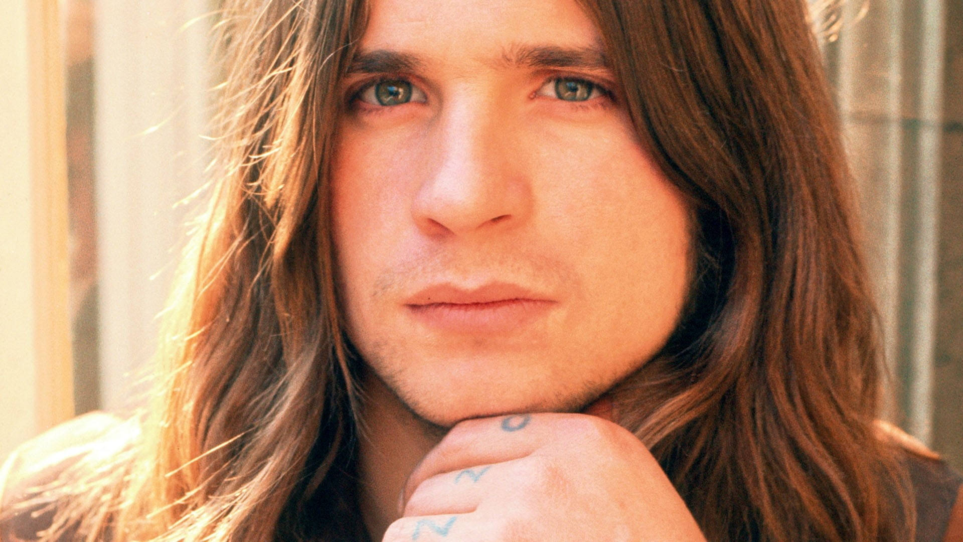Ozzy Osbourne 桌布 壁紙 — 一個長著棕色長髮和藍眼睛的男人，將下巴擱在刺青手上的特寫肖像。