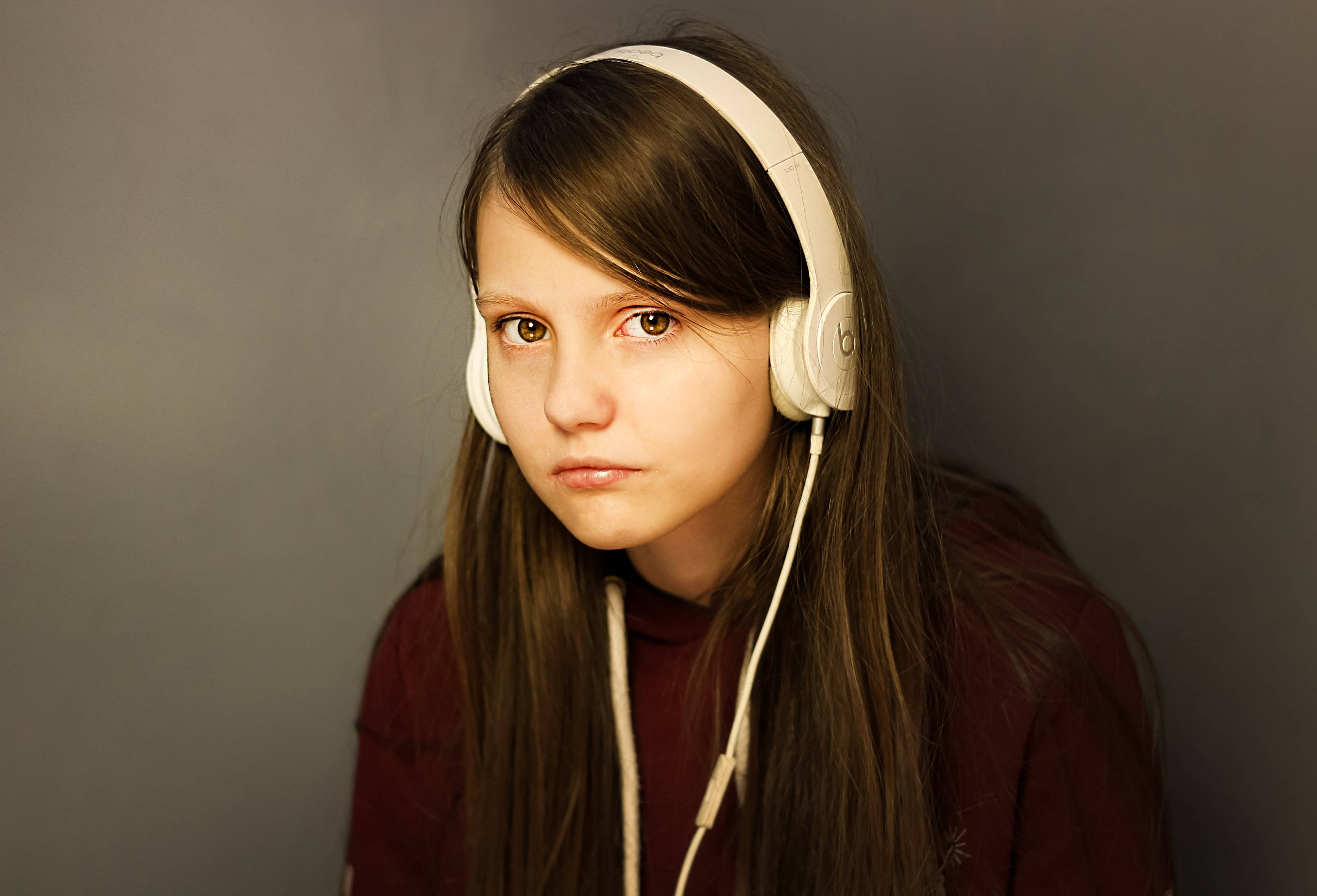 Teenage Girl 桌布 壁紙 — 一個長著棕色長髮的年輕女孩戴著白色耳機，在純灰色背景前專注地凝視著相機。
