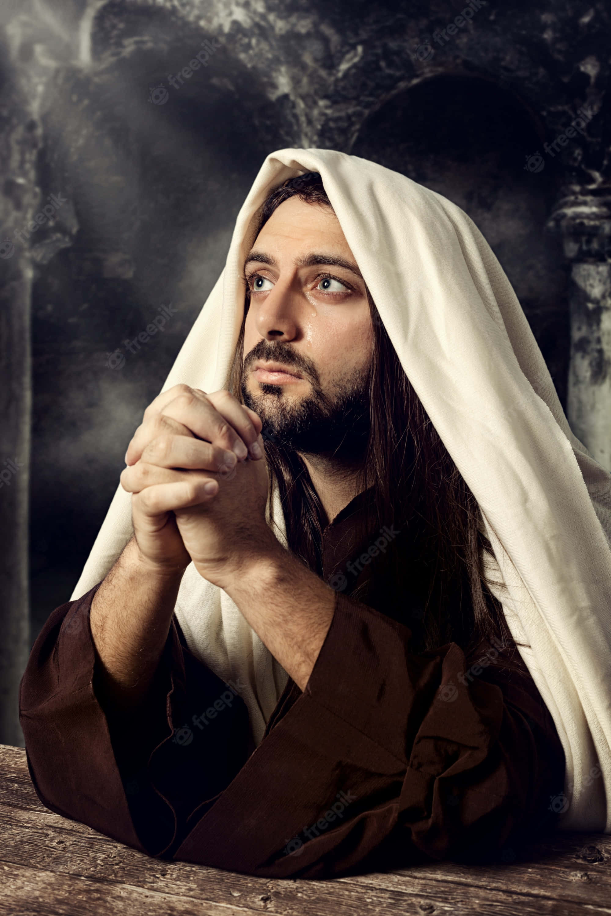 Jesus Praying 桌布 壁紙 — 一個長著深色長髮和鬍鬚的男人，披著白布，在黑暗的石拱門前合掌祈禱。