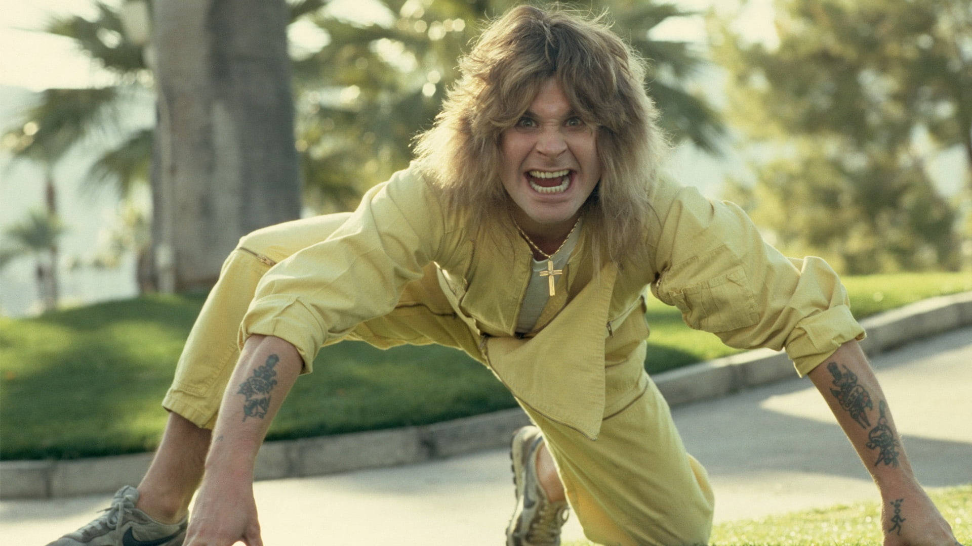 Ozzy Osbourne 桌布 壁紙 — 一個長金髮、有刺青的男人，穿著黃色連身褲，在陽光明媚的公園背景下咄咄逼人地蹲著。