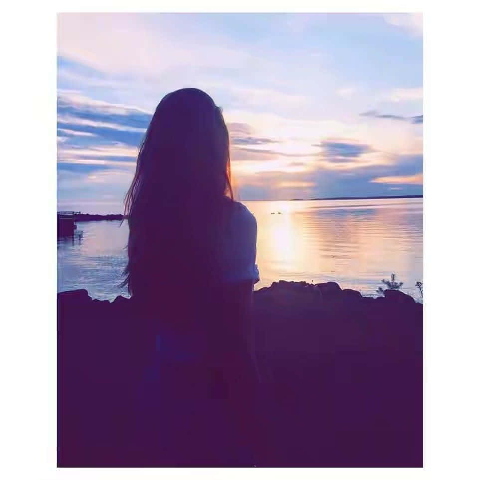 Instagram Profile 桌布 壁紙 — 一個長髮人物的剪影，凝視著平靜水面上柔和的紫色和橙色色調的寧靜日落。