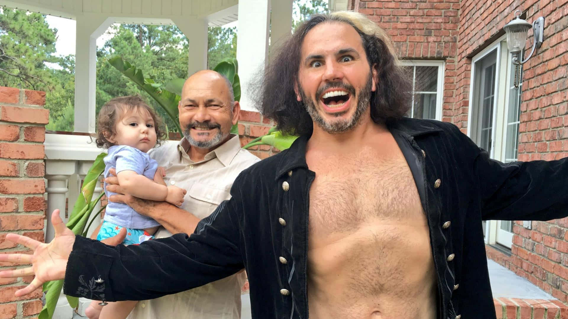 WWE 桌布 壁紙 — 一個長髮男人張開雙臂，抱著一個嬰兒，開心大笑，站在磚砌門廊上，旁邊另一個男人微笑著。