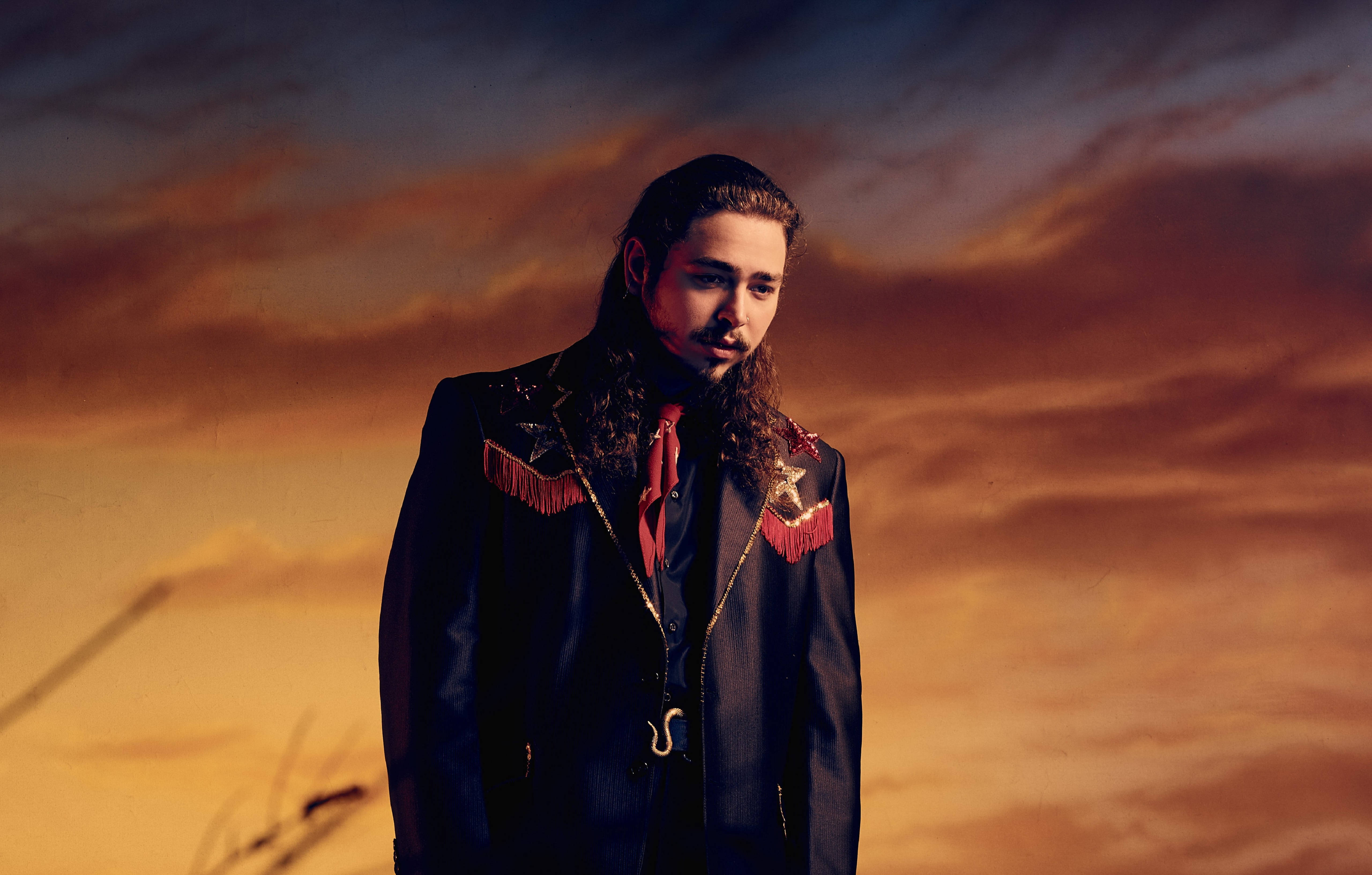 Post Malone 桌布 壁紙 — 一個長髮男子穿著流蘇黑色外套，站在戲劇性的日落天空前，天空有橙色和紫色的雲。