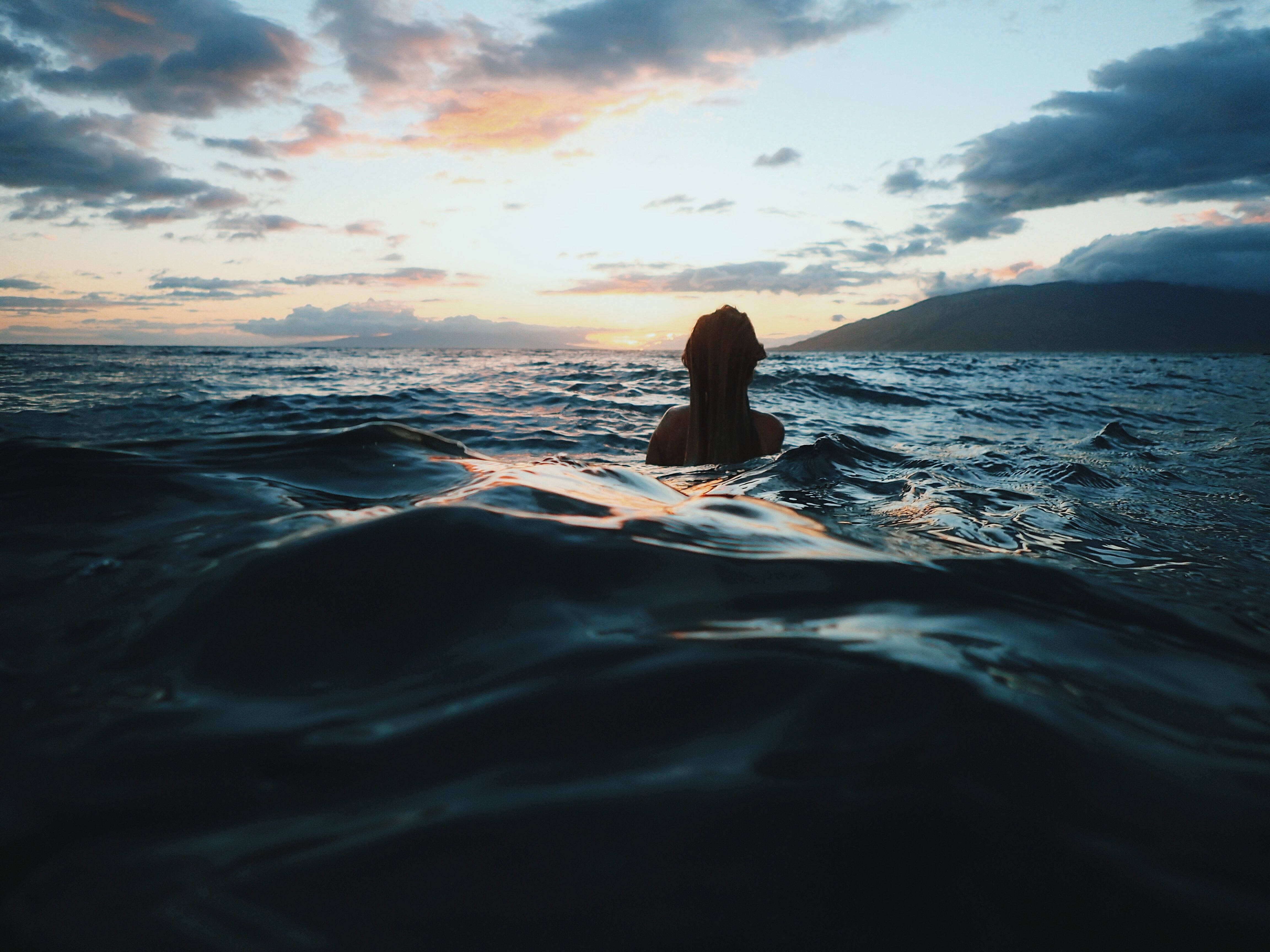 4K 沉靜 桌布 壁紙 — 一個長髮的人在海浪中平靜地漂浮，夕陽西下，遠處有山的剪影。