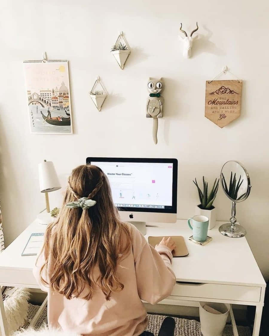 White Aesthetic Laptop 桌布 壁紙 — 一個長髮的人坐在白色書桌前使用電腦，周圍是裝飾性牆壁藝術和植物，在舒適的家庭辦公室中。