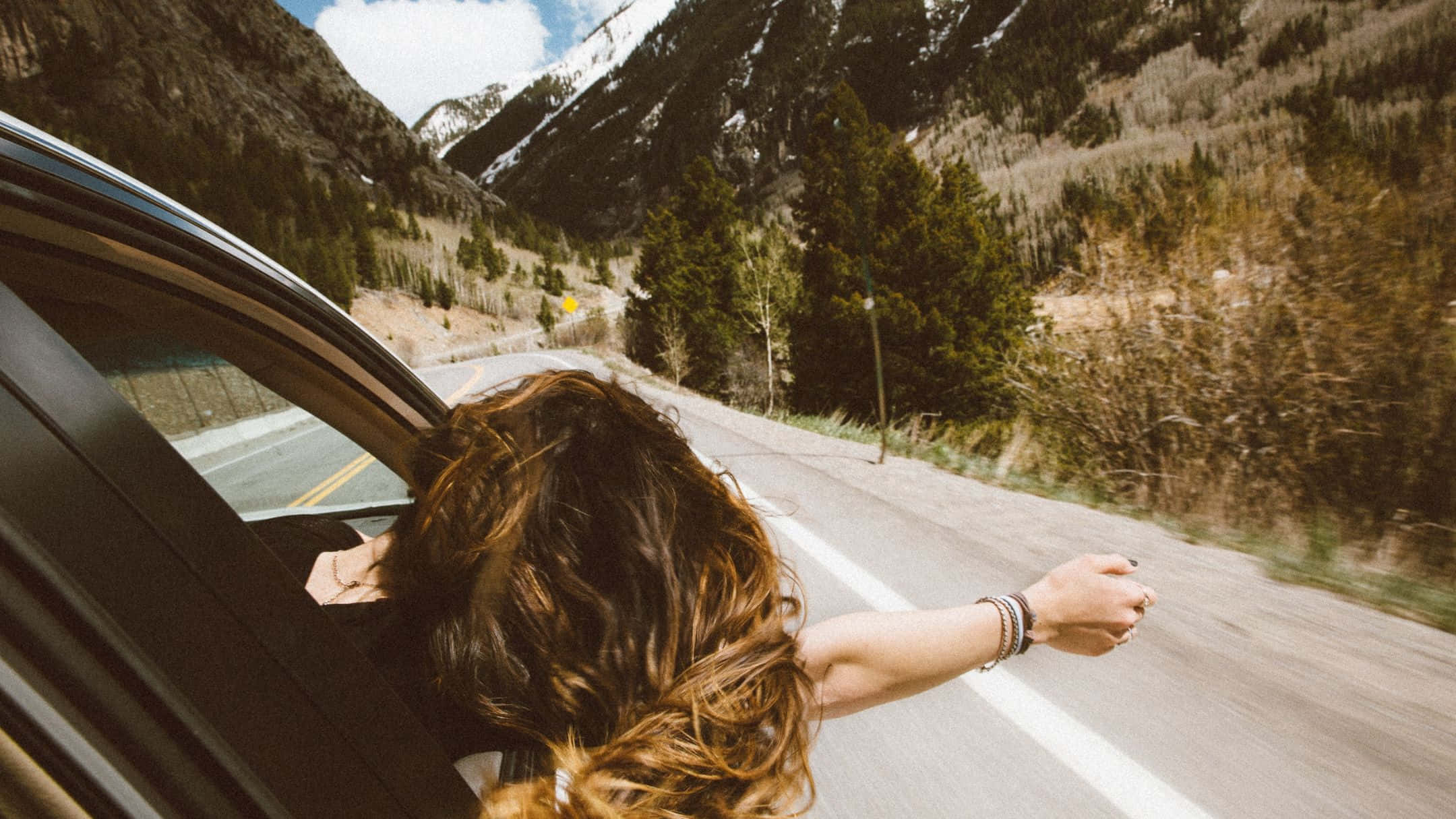 road trip 桌布 壁紙 — 一個長髮的人從行駛中的汽車車窗探出身子，雙臂張開，車輛沿著蜿蜒的山路行駛，兩旁是常綠森林和白雪覆蓋的山峰，天空明亮。