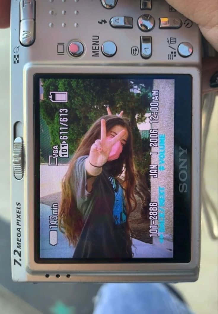 Holding Camera 桌布 壁紙 — 一個長髮的人戴著粉紅色口罩，在銀色Sony相機的LCD螢幕上被看到時做出和平手勢。