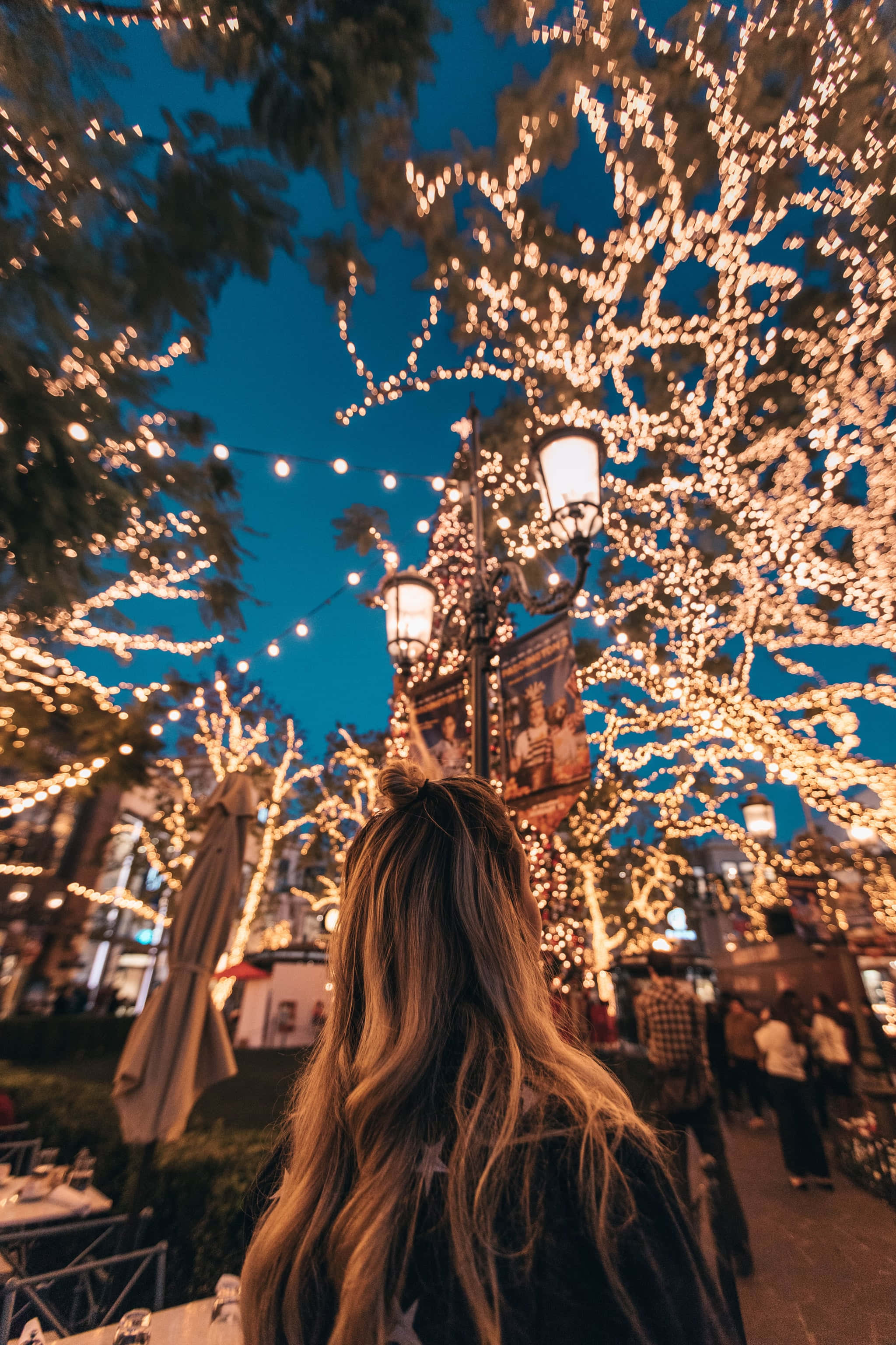 iPhone Christmas Aesthetic 桌布 壁紙 — 一個長髮的人抬頭仰望被發光燈串纏繞的樹木，背景是深藍色的夜空。