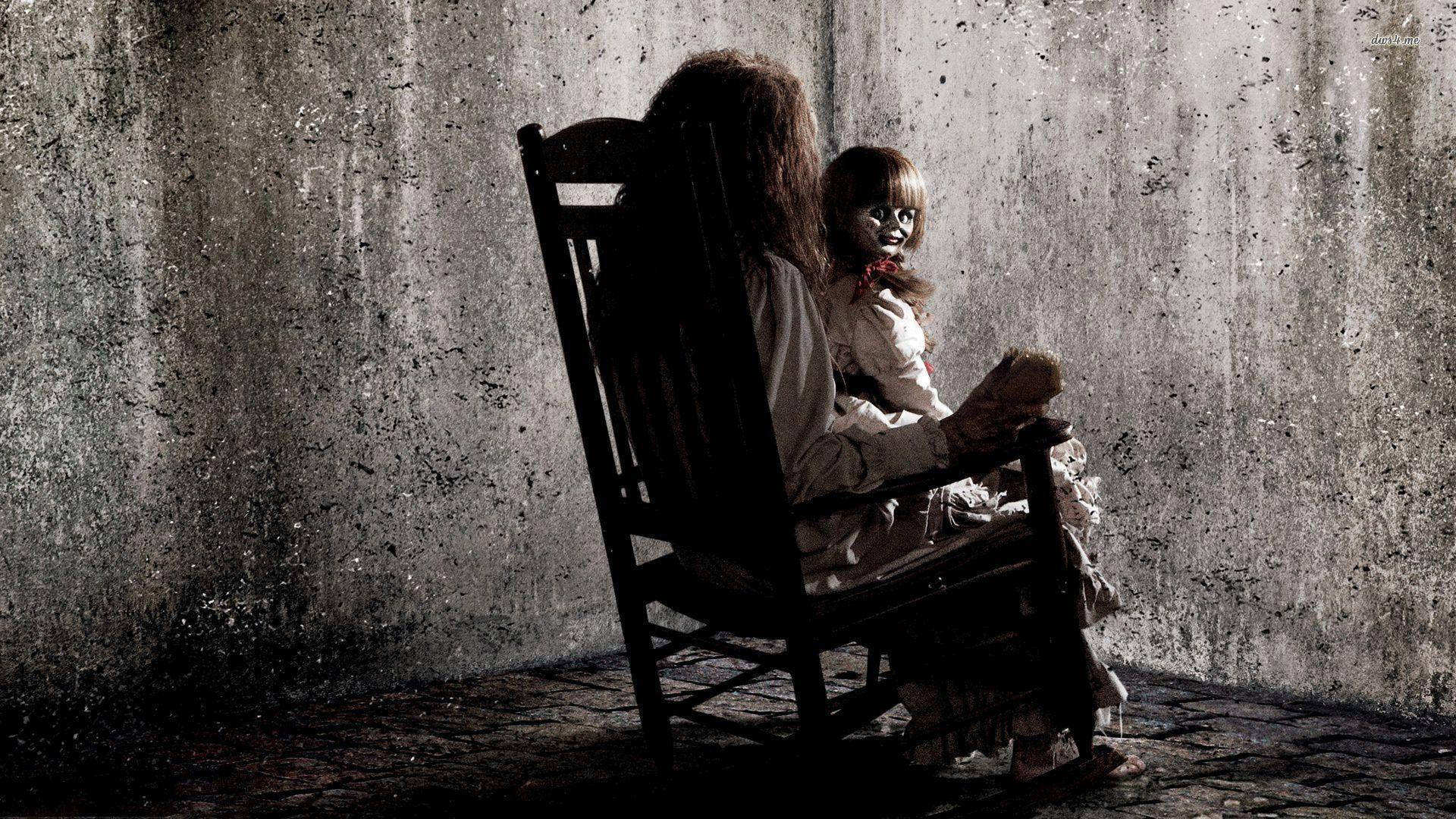 Annabelle 桌布 壁紙 — 一個長髮的恐怖人物坐在搖椅上，抱著一個臉色蒼白、嘴唇鮮紅的娃娃，背景是污穢的混凝土牆。