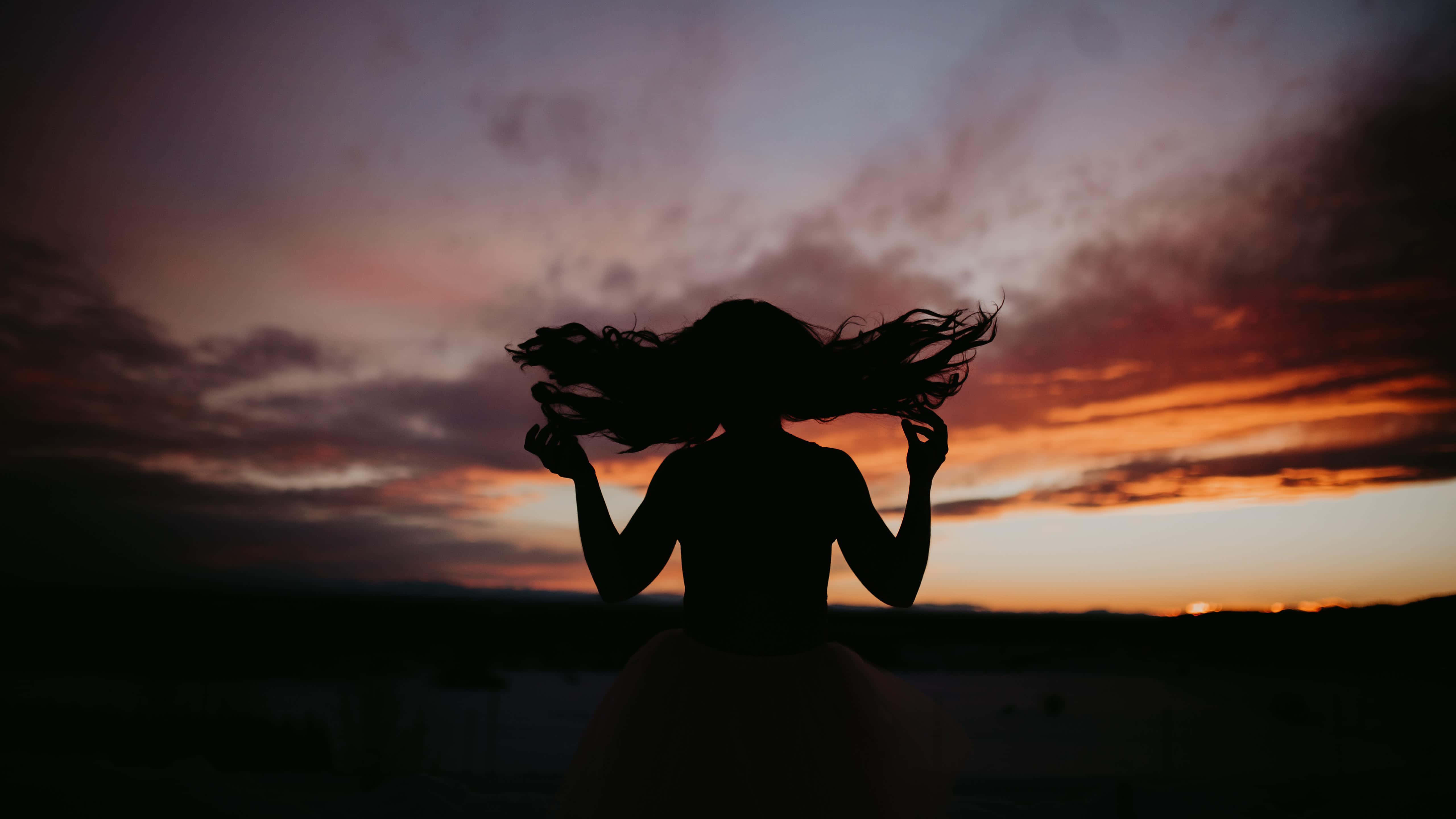 Sunset Silhouette 桌布 壁紙 — 一個長髮飄逸的女人的剪影，映襯著充滿活力的日落天空，有紫色、橙色和粉紅色的雲彩。