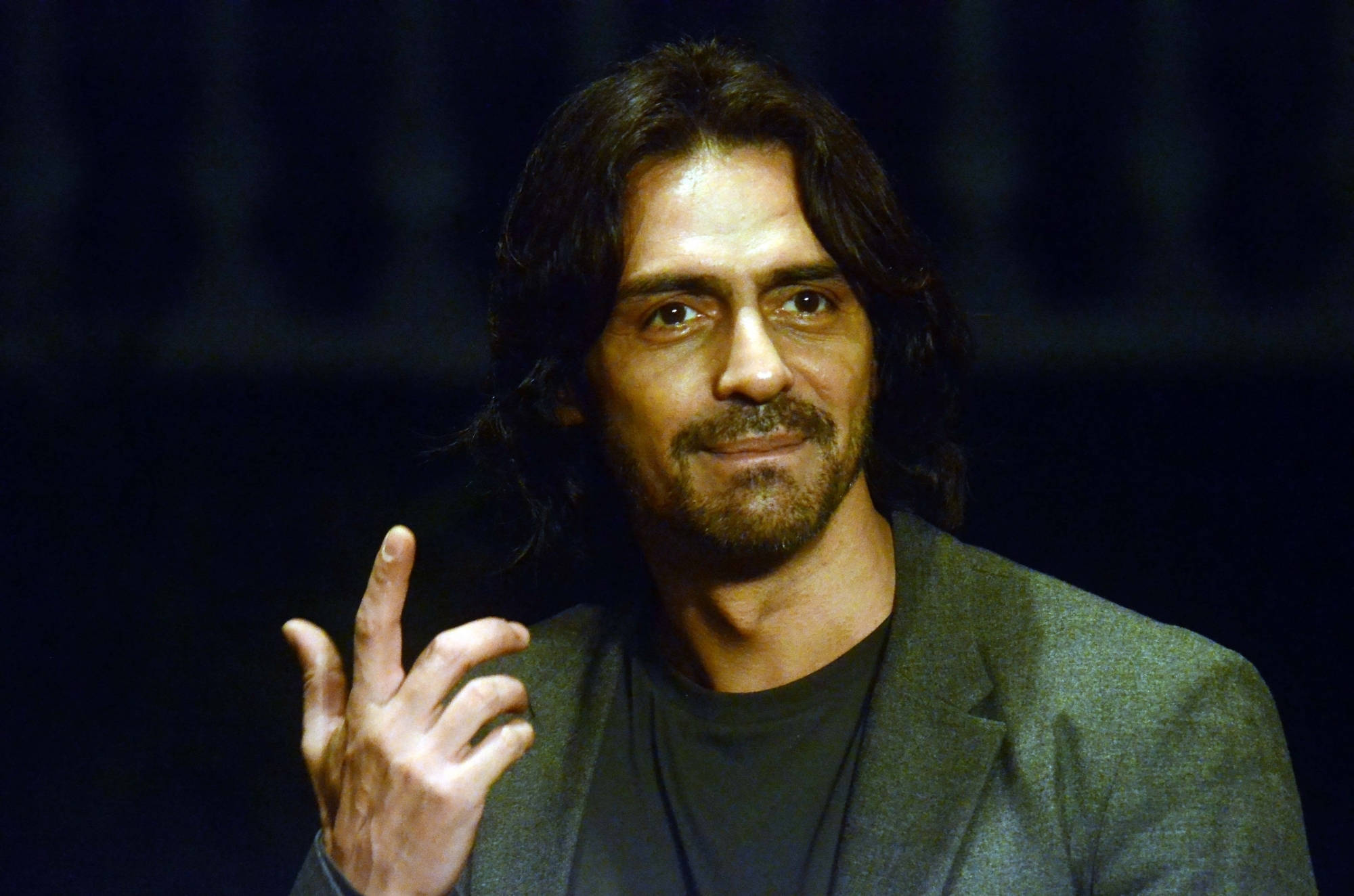 Arjun Rampal 桌布 壁紙 — 一個長黑髮、留鬍鬚的男人，身穿灰色西裝外套配黑色襯衫，在深色背景前做手勢。