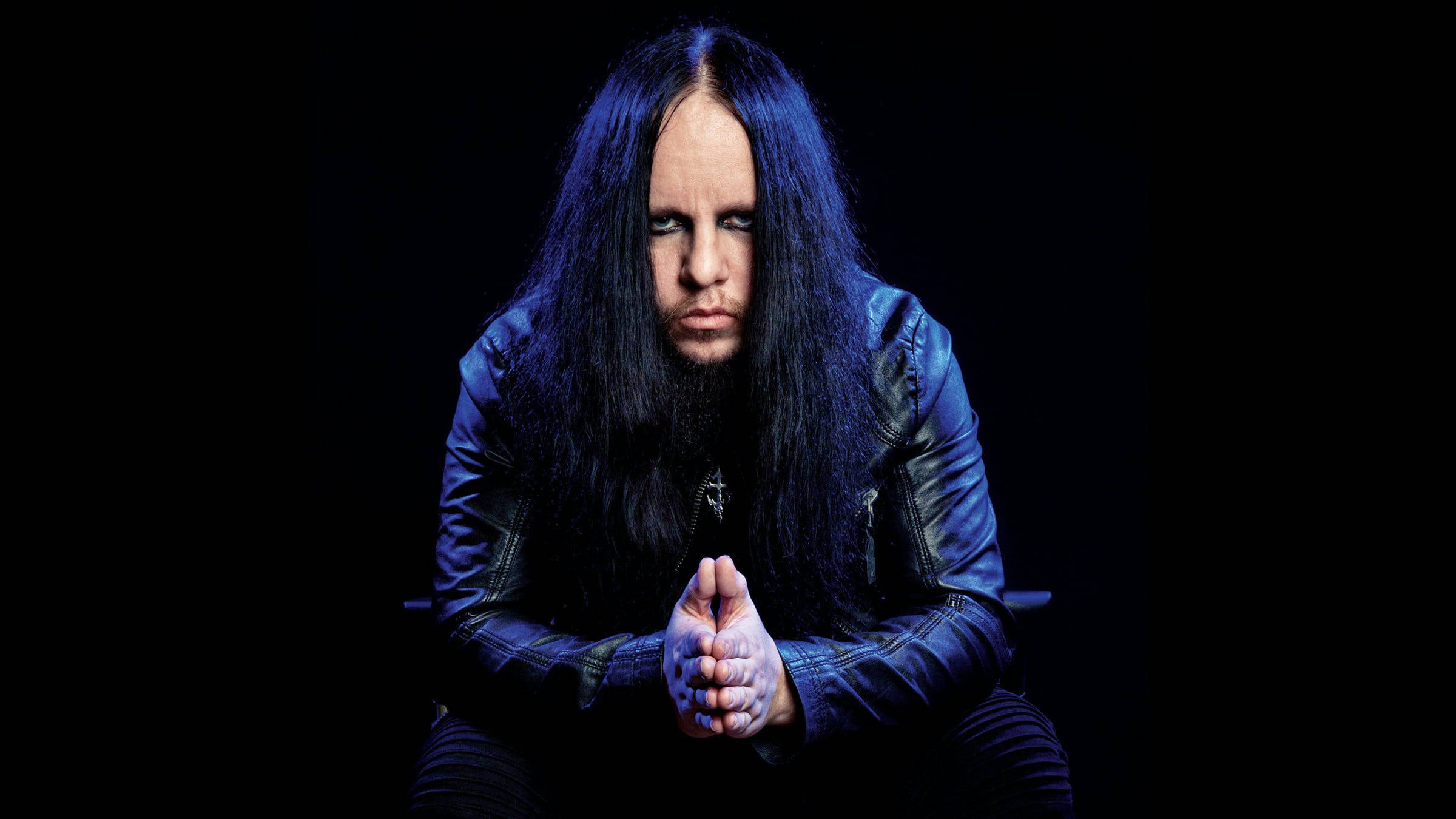 Joey Jordison 桌布 壁紙 — 一個長黑髮、眼神銳利的男人，穿著皮夾克，坐在黑色背景前，帶有戲劇性的藍色燈光。