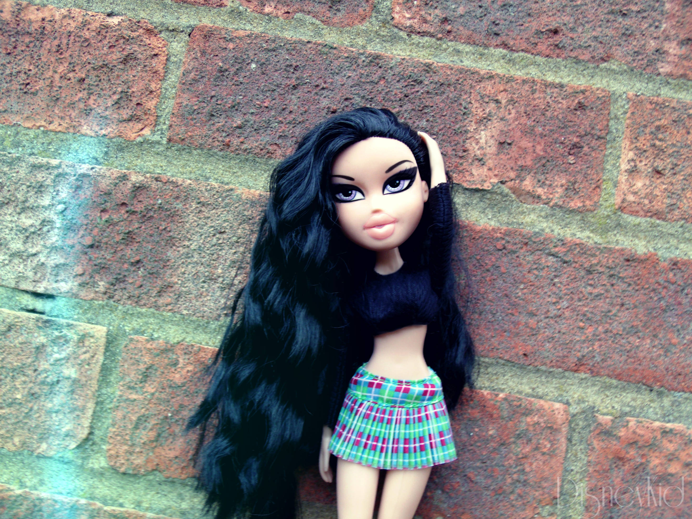 bratz 美學 桌布 壁紙 — 一個長黑髮、穿著格子裙的 Bratz 公仔靠在一面帶有綠色塗鴉的風化磚牆上。