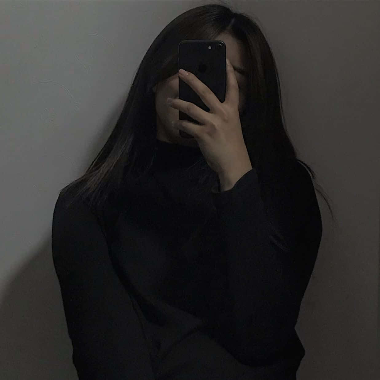 Dating Profile 桌布 壁紙 — 一個長黑髮的人拿著電話遮住臉，靠著素牆，光線昏暗。