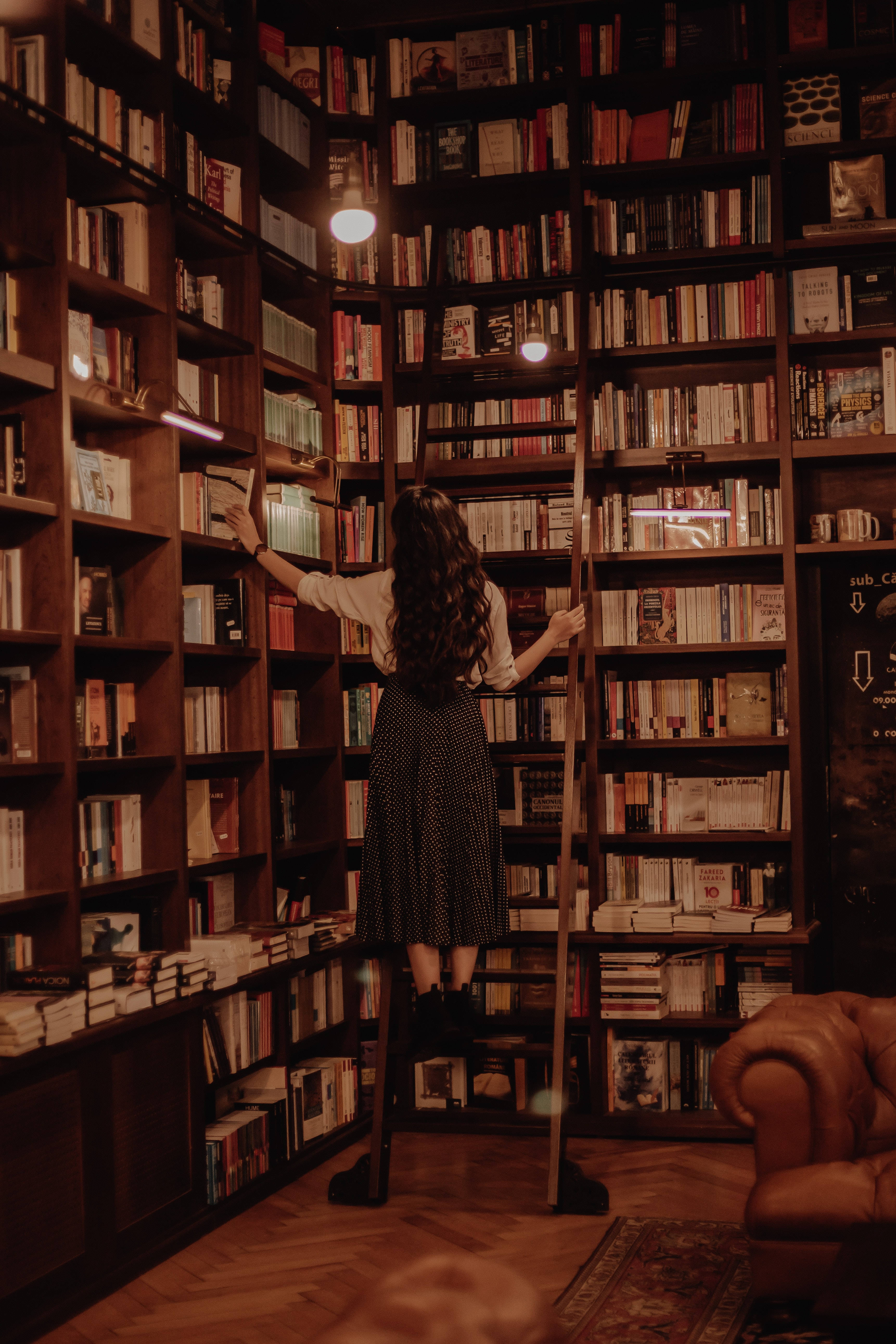Library 桌布 壁紙 — 一個長黑髮的人站在梯子上，伸手去拿一個溫暖照明的木質圖書館裡高聳書架上的書。