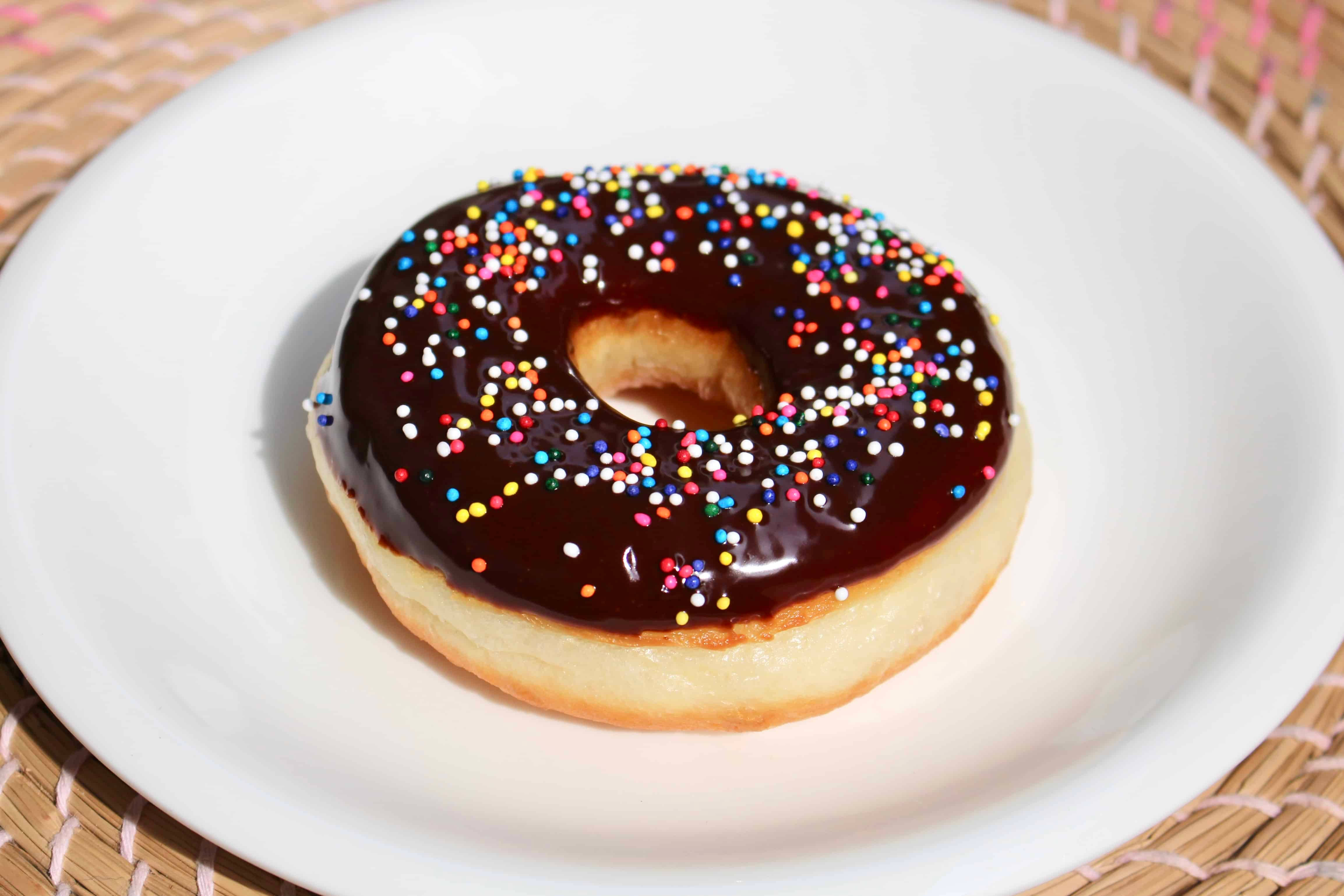 Donut 桌布 壁紙 — 一個閃亮的巧克力糖霜甜甜圈，上面有彩色糖粒，放在白色盤子上，置於編織竹蓆上。