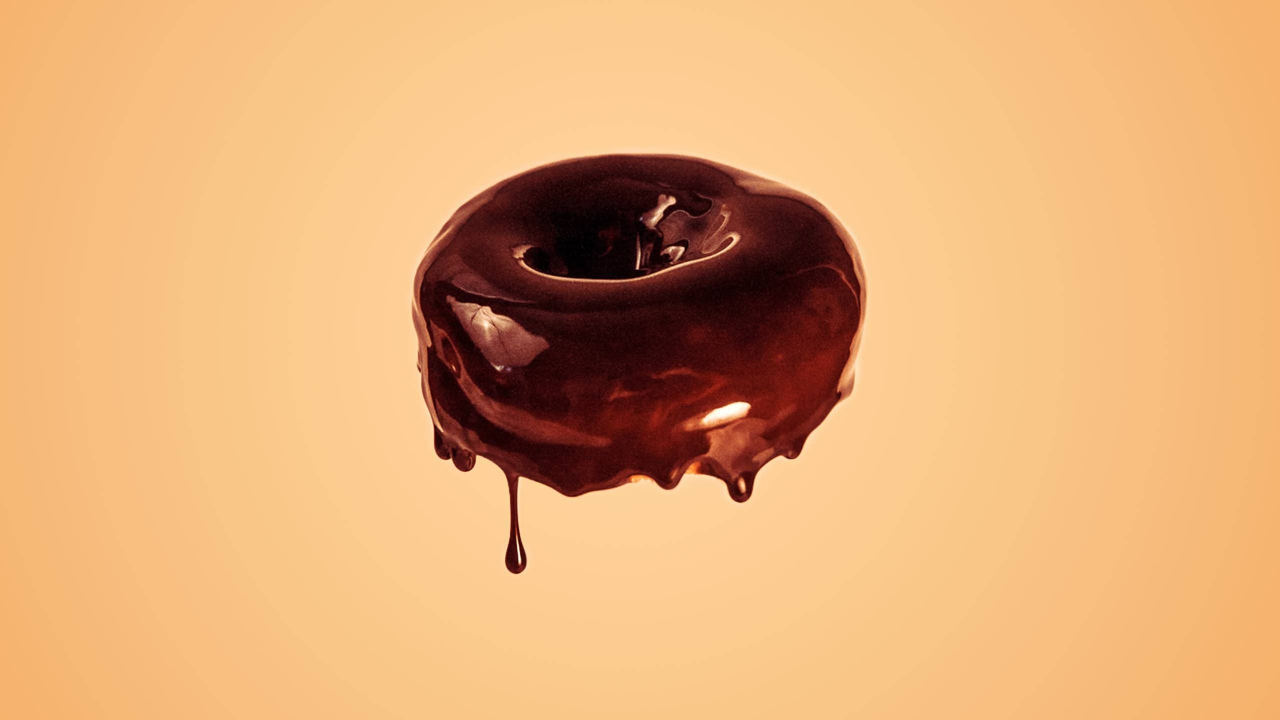 Donut 桌布 壁紙 — 一個閃亮的巧克力釉面甜甜圈，滴落的糖霜漂浮在溫暖的蜜桃色背景上。