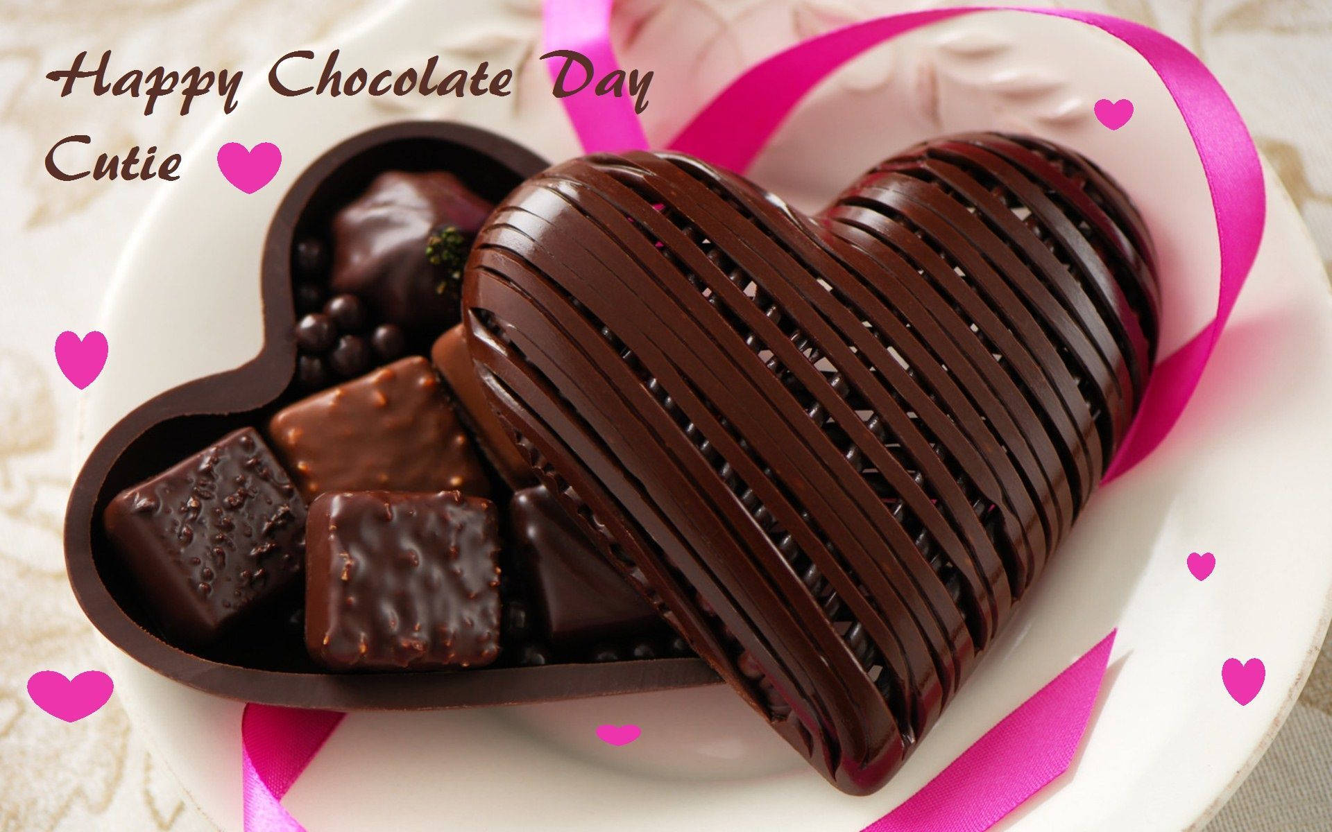 Chocolate 桌布 壁紙 — 一個閃亮的心形巧克力盒，裡面裝滿了各種巧克力，放在一個白色盤子上，配有粉紅色絲帶和心形裝飾。