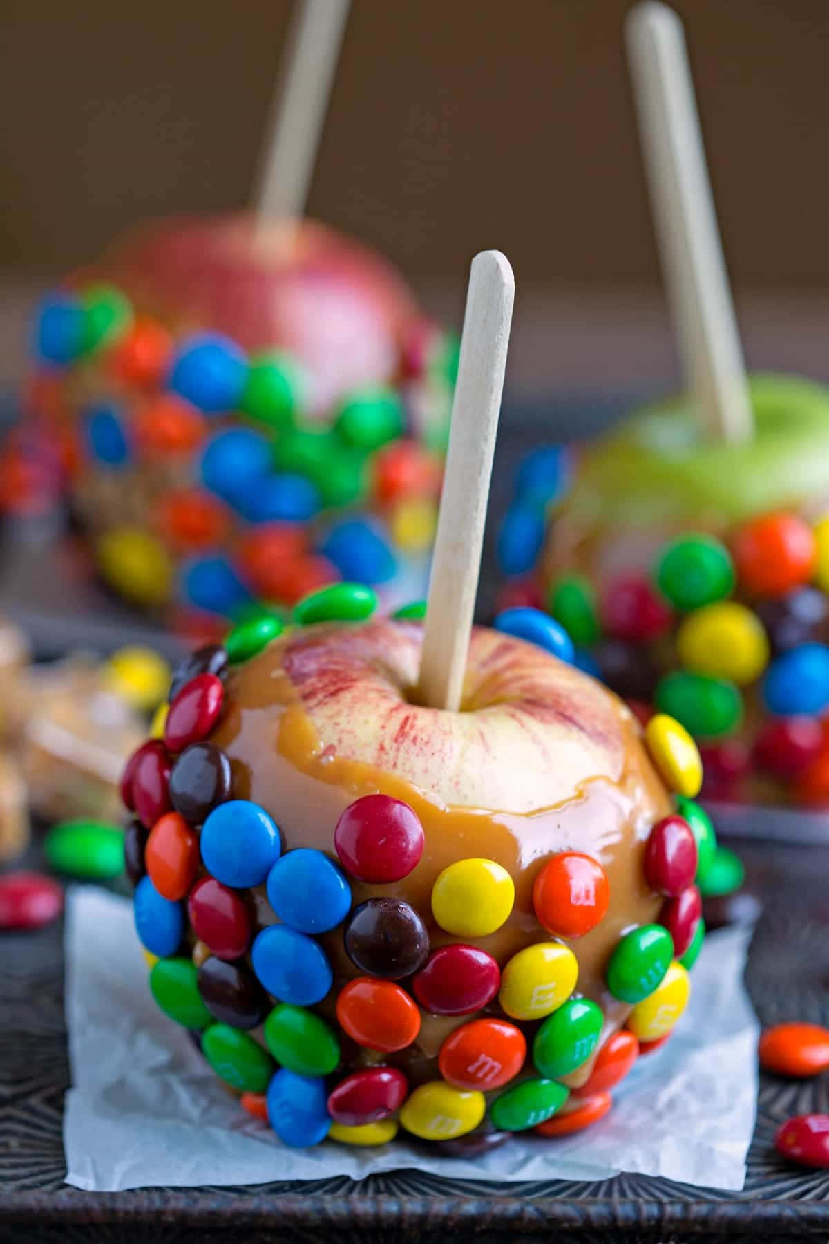 Caramel Apples 桌布 壁紙 — 一個閃亮的焦糖蘋果，上面裝飾著色彩繽紛的M&M's糖果，放在深色木質表面上。