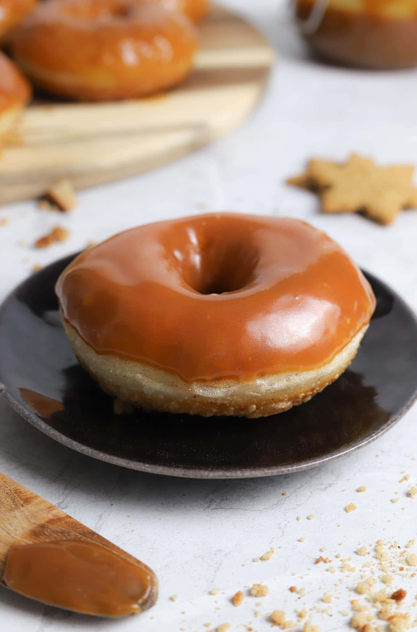 Glazed Donut 桌布 壁紙 — 一個閃亮的焦糖釉面甜甜圈放在深色盤子上，旁邊有散落的碎屑和星形餅乾。