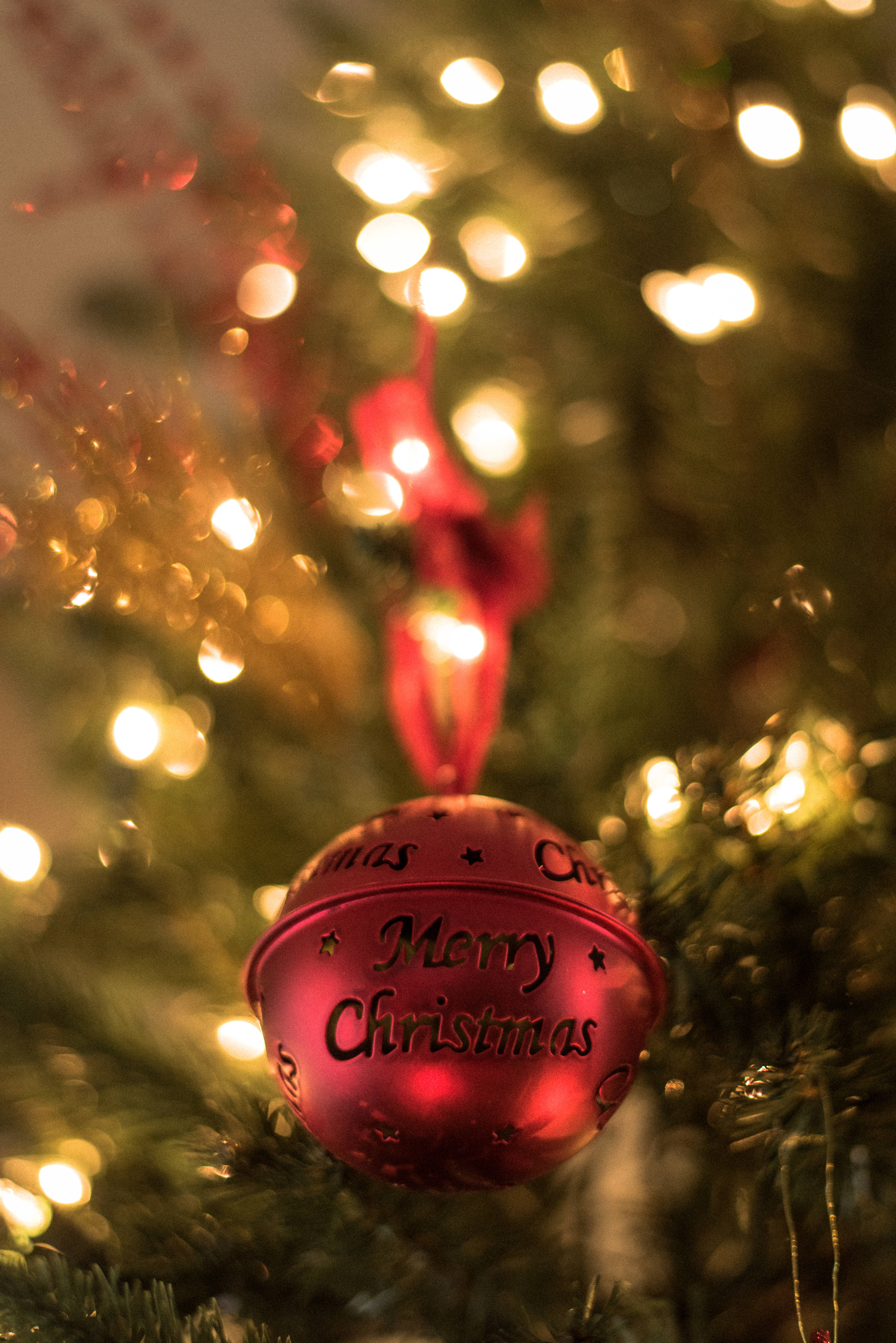 聖誕節 桌布 壁紙 — 一個閃亮的紅色聖誕裝飾，上面有「Merry Christmas」字樣，掛在裝飾著閃爍仙女燈的松樹上。