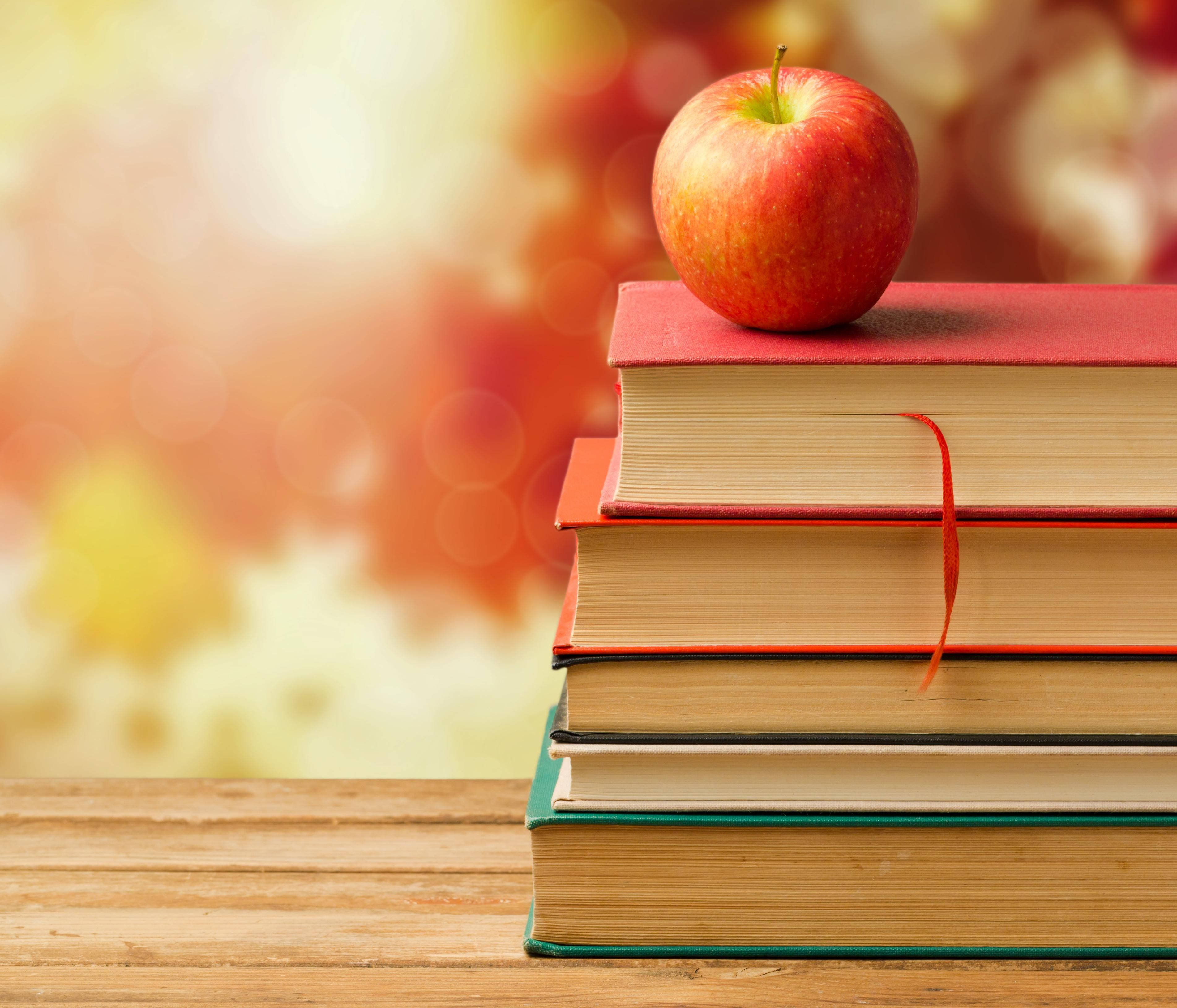 學校 桌布 壁紙 — 一個閃亮的紅蘋果放在一堆色彩繽紛的書本上，置於木質表面，背景是溫暖的散景效果。