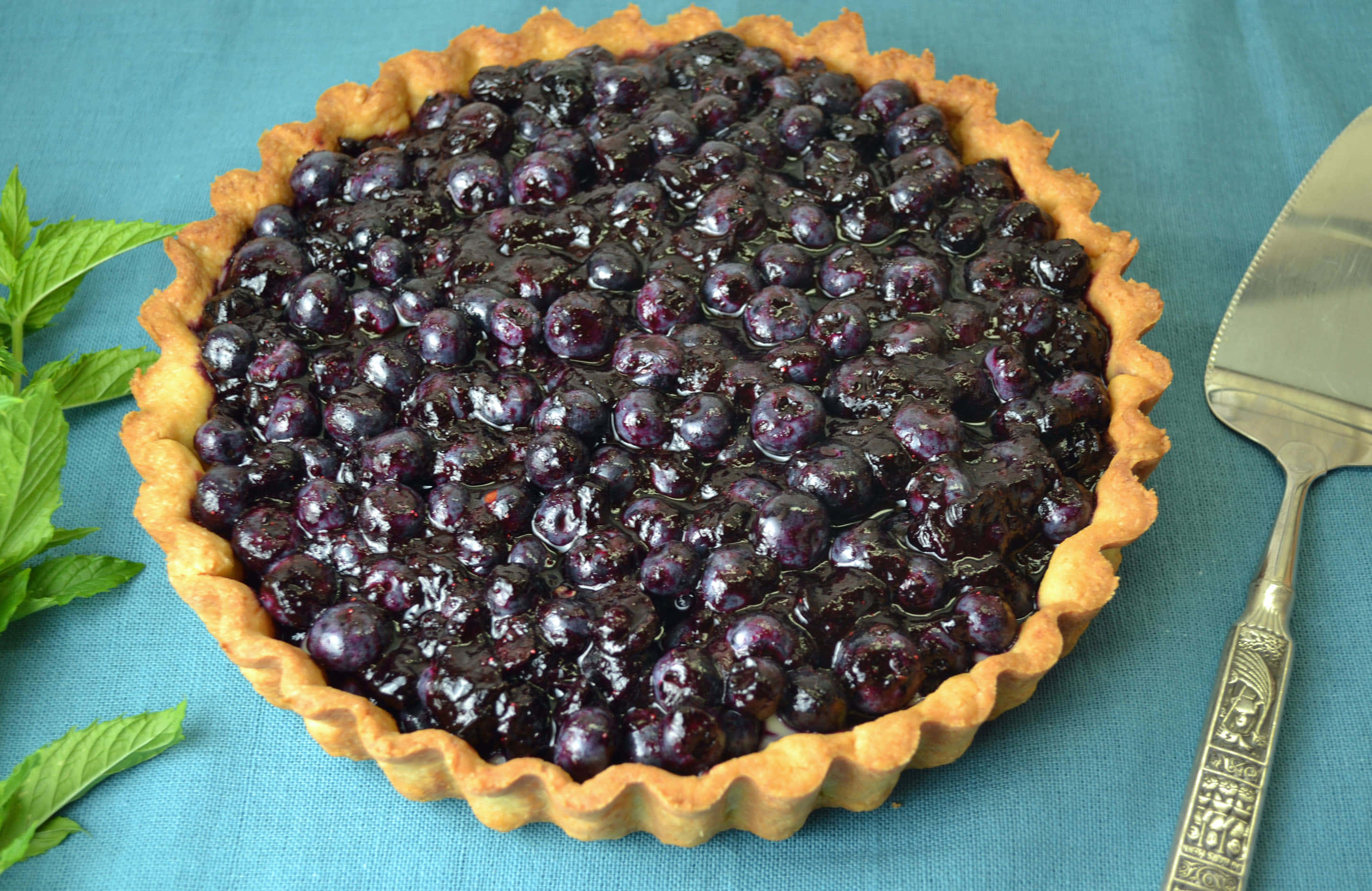 Blueberries Tart 桌布 壁紙 — 一個閃亮的藍莓塔，金黃色的塔皮放在青綠色布料上，旁邊有薄荷葉和裝飾用服務鏟。