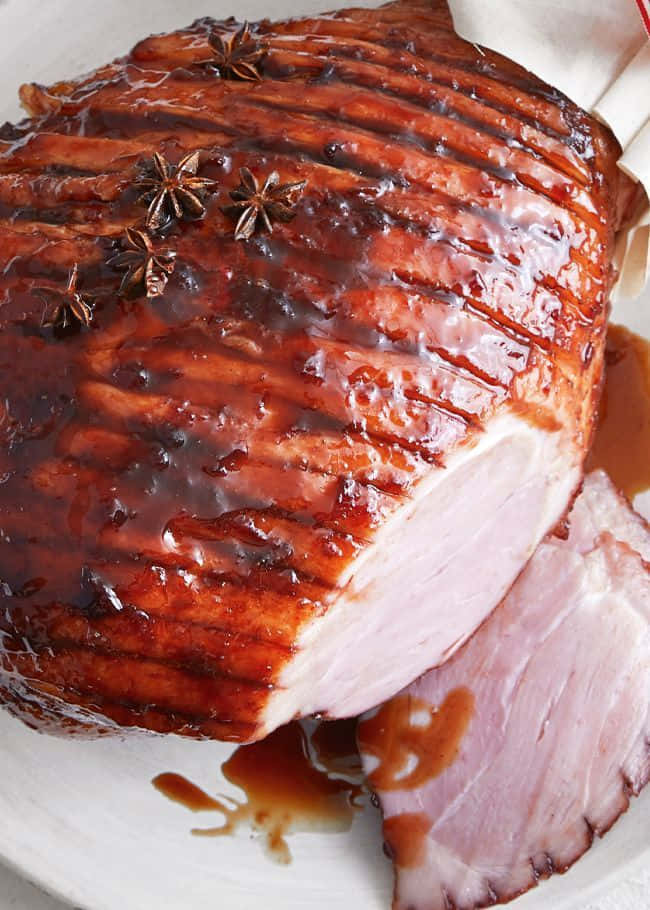 Black Forest Ham 桌布 壁紙 — 一個閃亮的蜜汁火腿，上面點綴著八角，放在白色盤子上，切開一片露出粉嫩的肉。