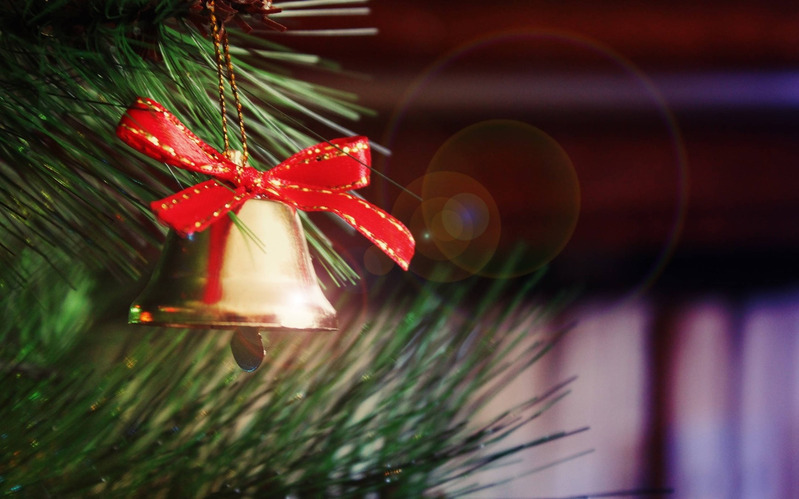 Christmas Bell 桌布 壁紙 — 一個閃亮的金色聖誕鈴鐺，飾有紅色絲帶，掛在綠色的松樹枝上，背景有溫暖的鏡頭光暈。